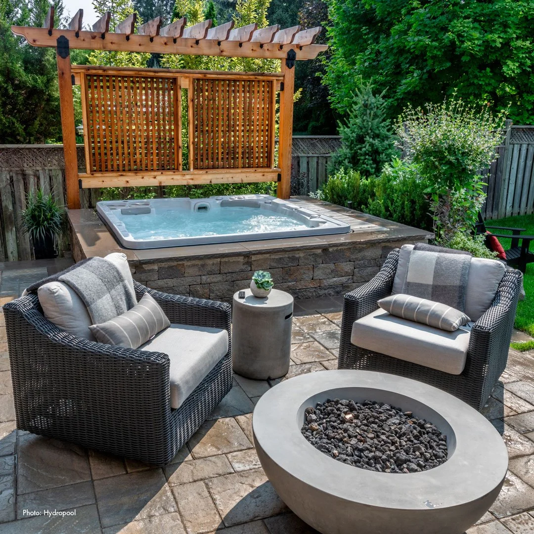 An outdoor patio with two cushioned wicker chairs, a small round side table, and a fire pit. Behind the seating area is a hot tub on a stone platform, partially enclosed by a wooden privacy screen, with lush green trees and shrubs surrounding the area.