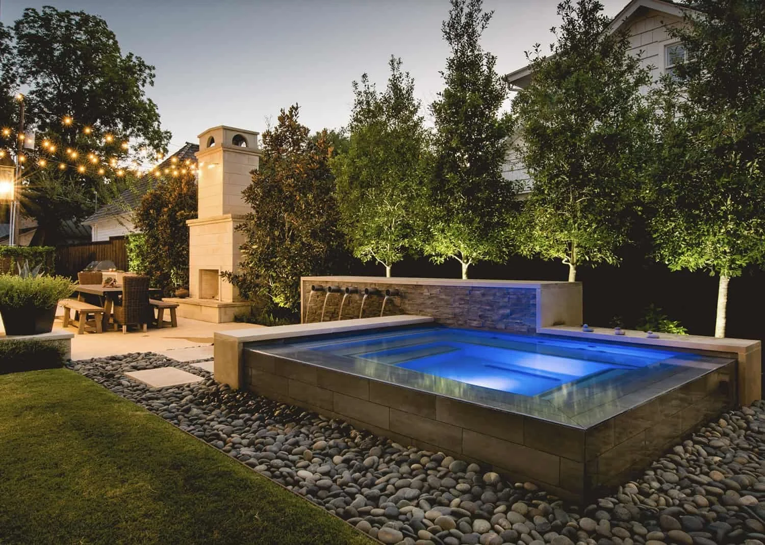 Backyard scene at dusk featuring a small illuminated hot tub surrounded by smooth rocks, a fire pit with seating, a fireplace with a chimney, string lights overhead, tall green trees, and a house in the background.
