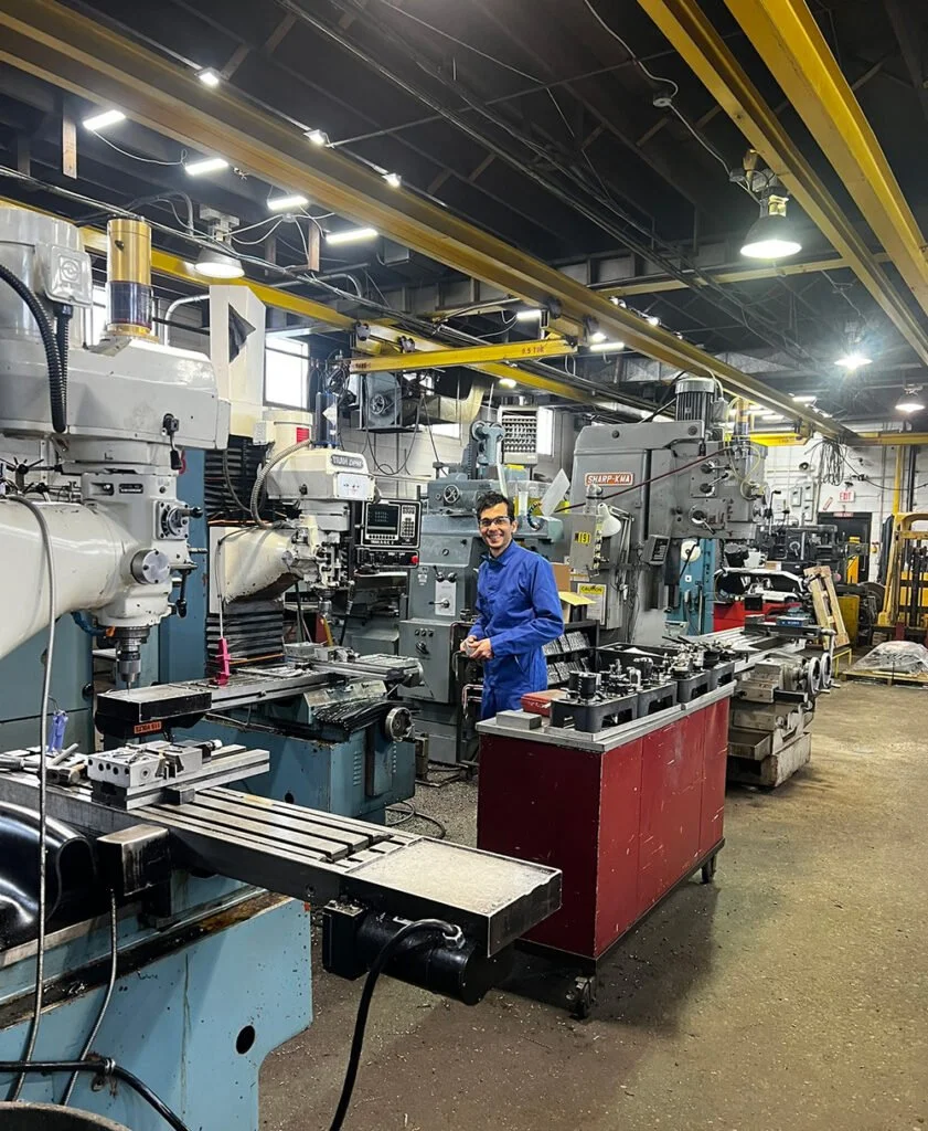 A person in a blue jumpsuit working in a machine shop with industrial milling machines and equipment.