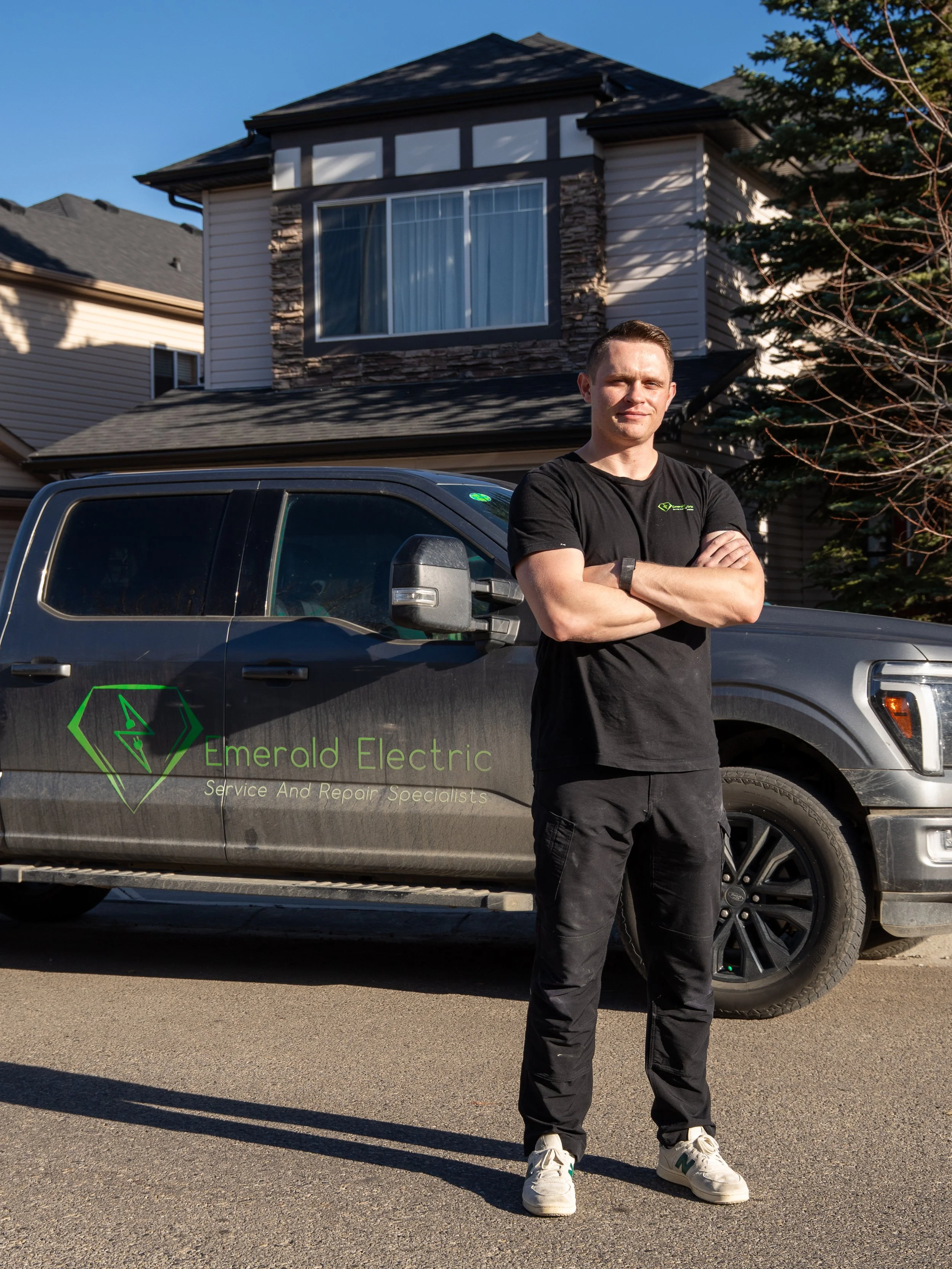 A man stands with arms crossed in front of a black pickup truck with the logo and name 'Emerald Electric' on the side, in a residential neighborhood with houses and trees in the background.