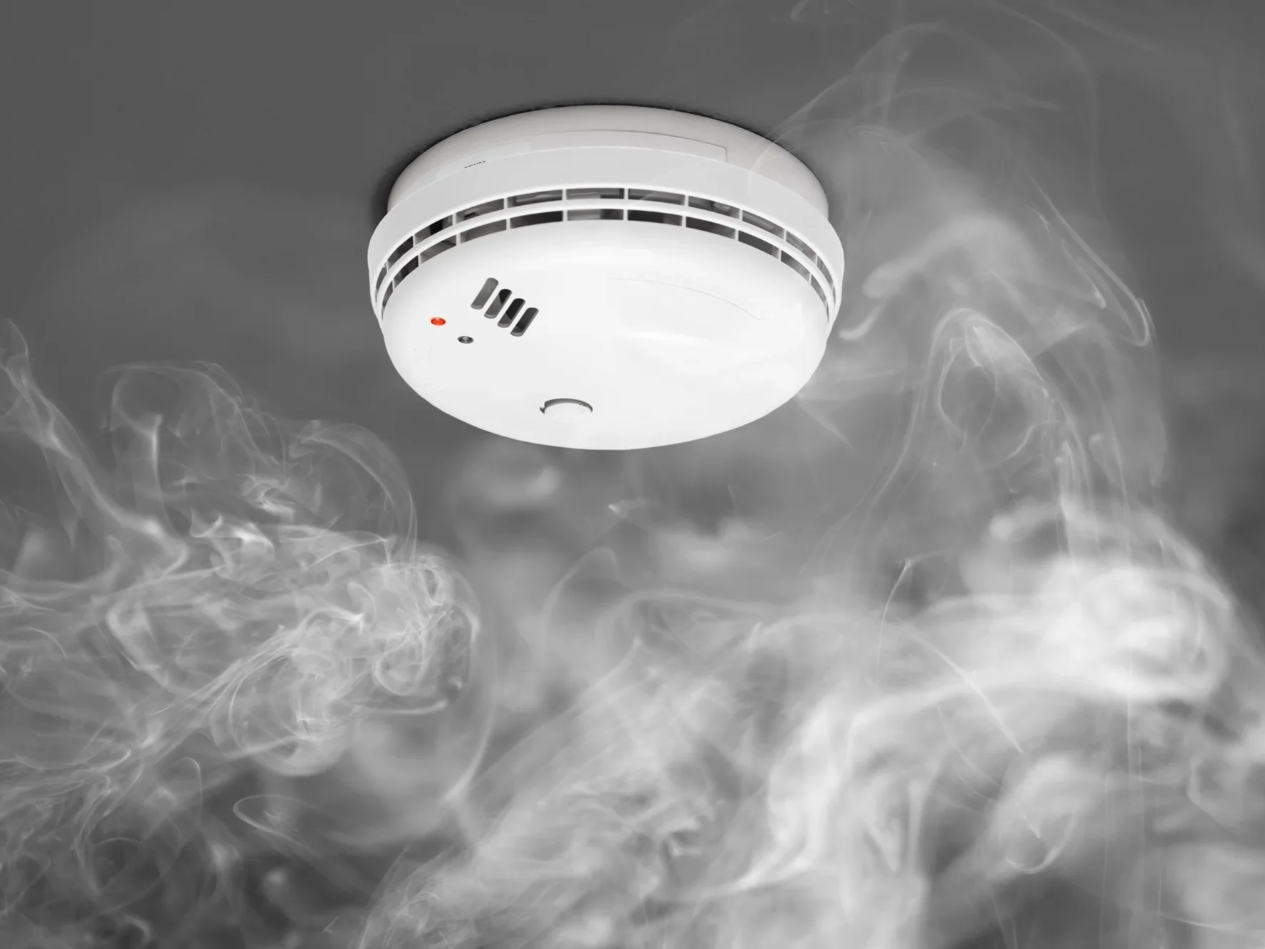 A smoke detector on the ceiling with smoke and wisps of smoke below it.