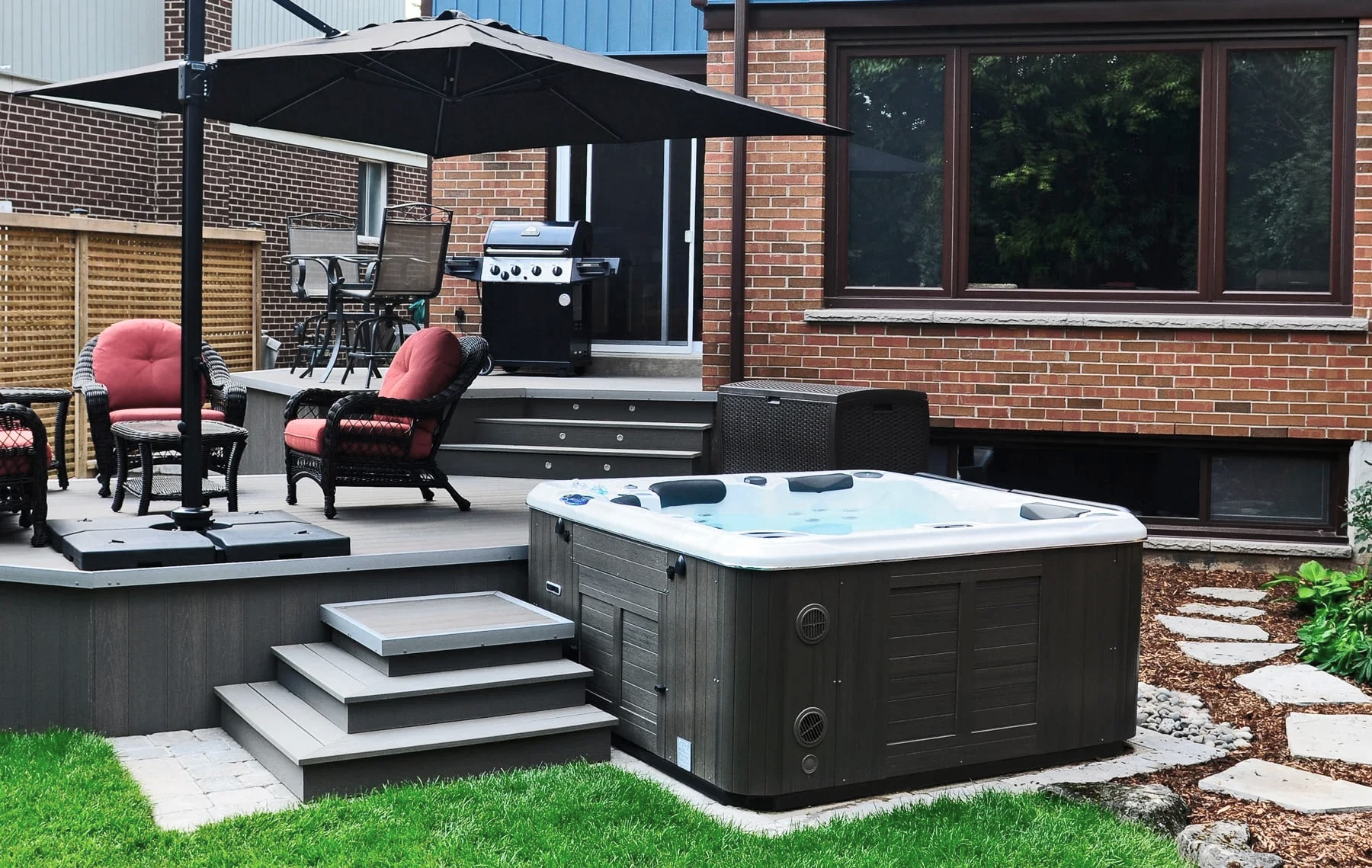Backyard deck with patio furniture, an umbrella, a grill, and a hot tub next to a brick house.