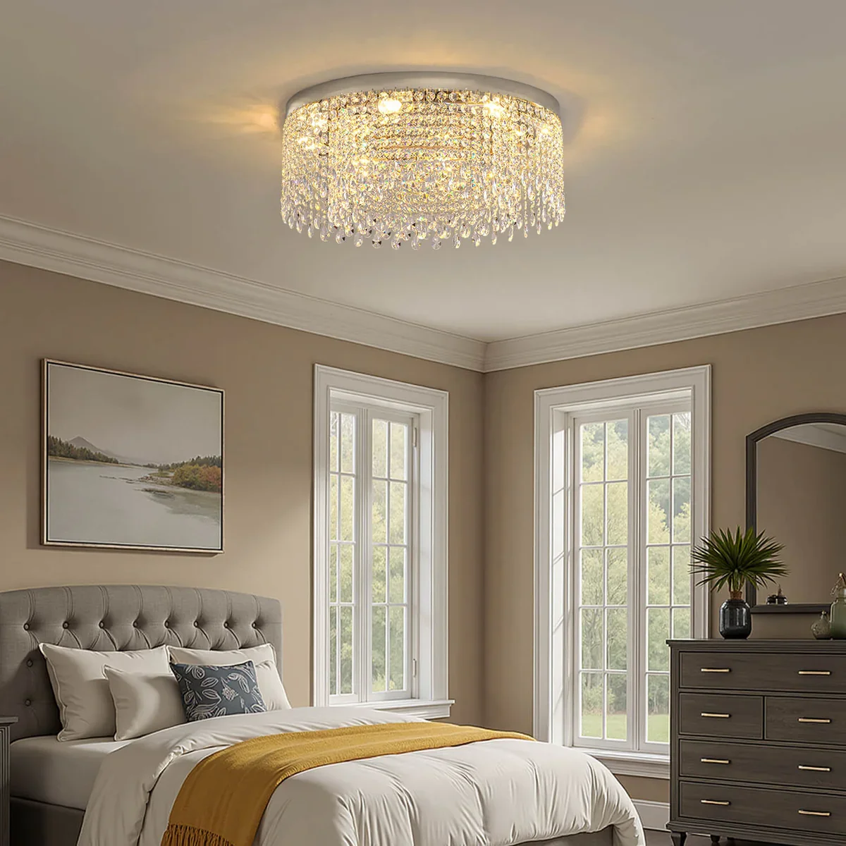 A bedroom with a crystal chandelier overhead, beige walls, two large windows, a gray upholstered bed with white and patterned pillows, a mustard yellow throw blanket, and a gray dresser with a plant and decorative items.