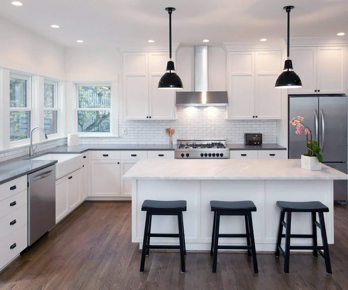 Modern white kitchen with gray countertops, black hardware, stainless steel appliances, a kitchen island with three black bar stools, large windows, a white subway tile backsplash, black pendant lights, and a potted orchid plant.