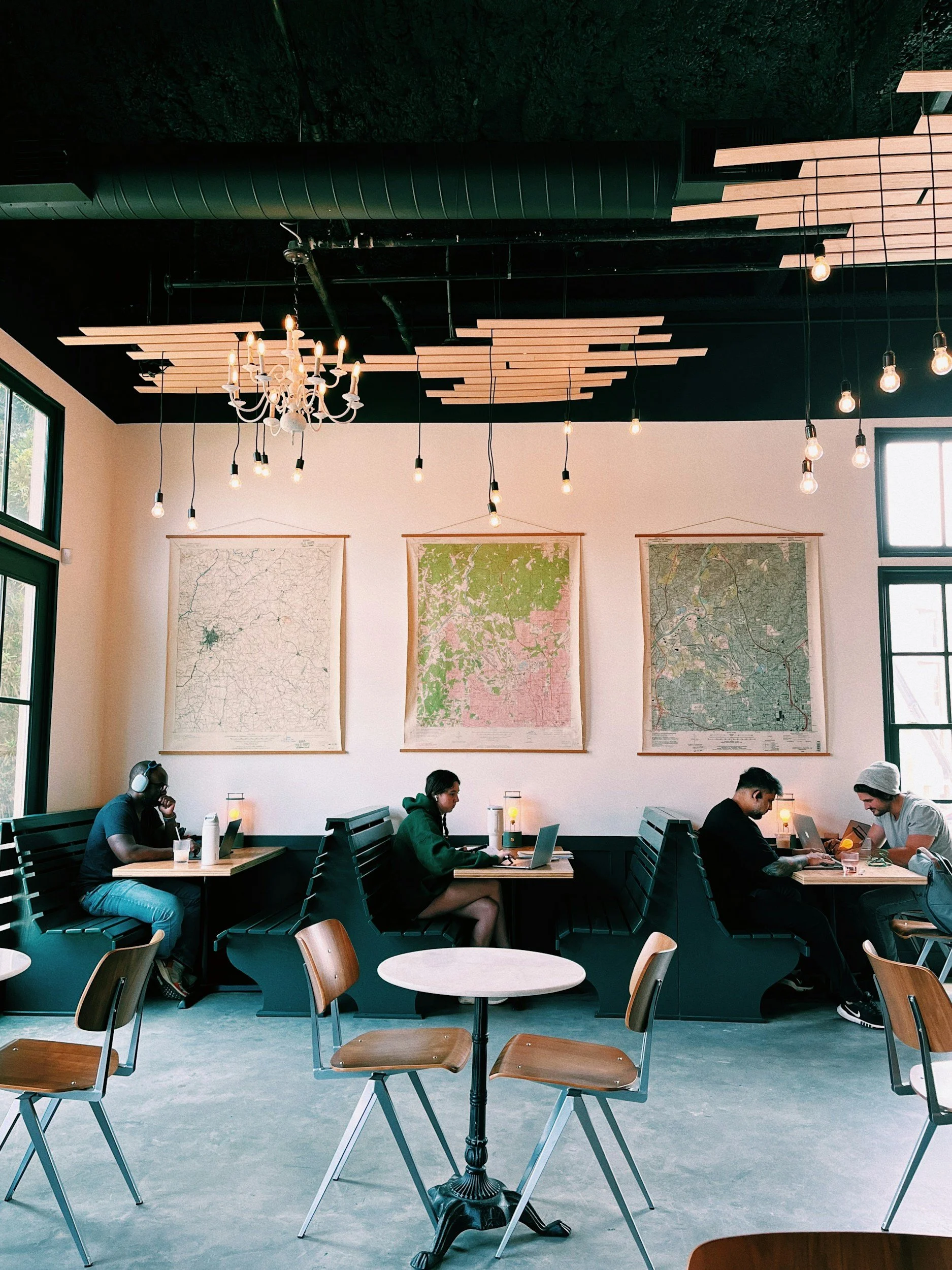 People working on laptops at tables in a modern coffee shop or café with large windows, wooden chairs, hanging light bulbs, and pictures of maps on the wall.