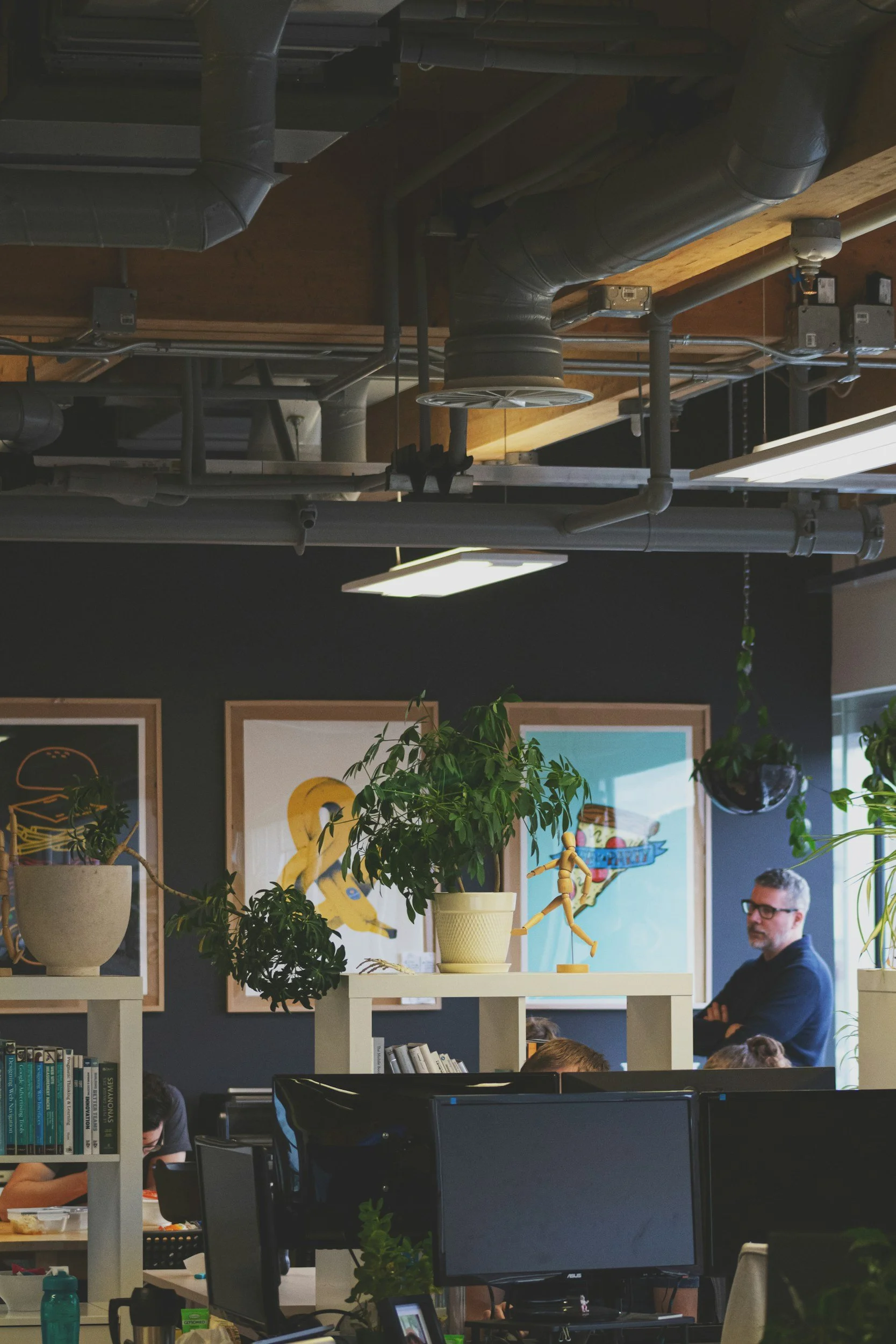 Office space with plants, artwork, and people working at desks, framed pictures on dark walls, and an open ceiling with exposed ductwork and lighting.