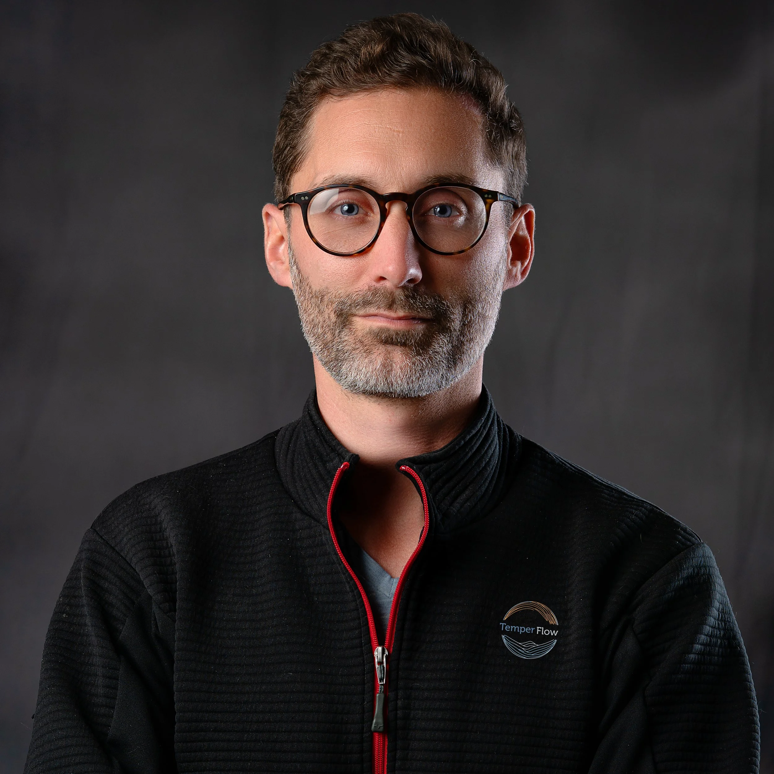 A man with dark hair, glasses, and a beard, wearing a dark sweater, looking straight ahead with a slight smile in an indoor setting.