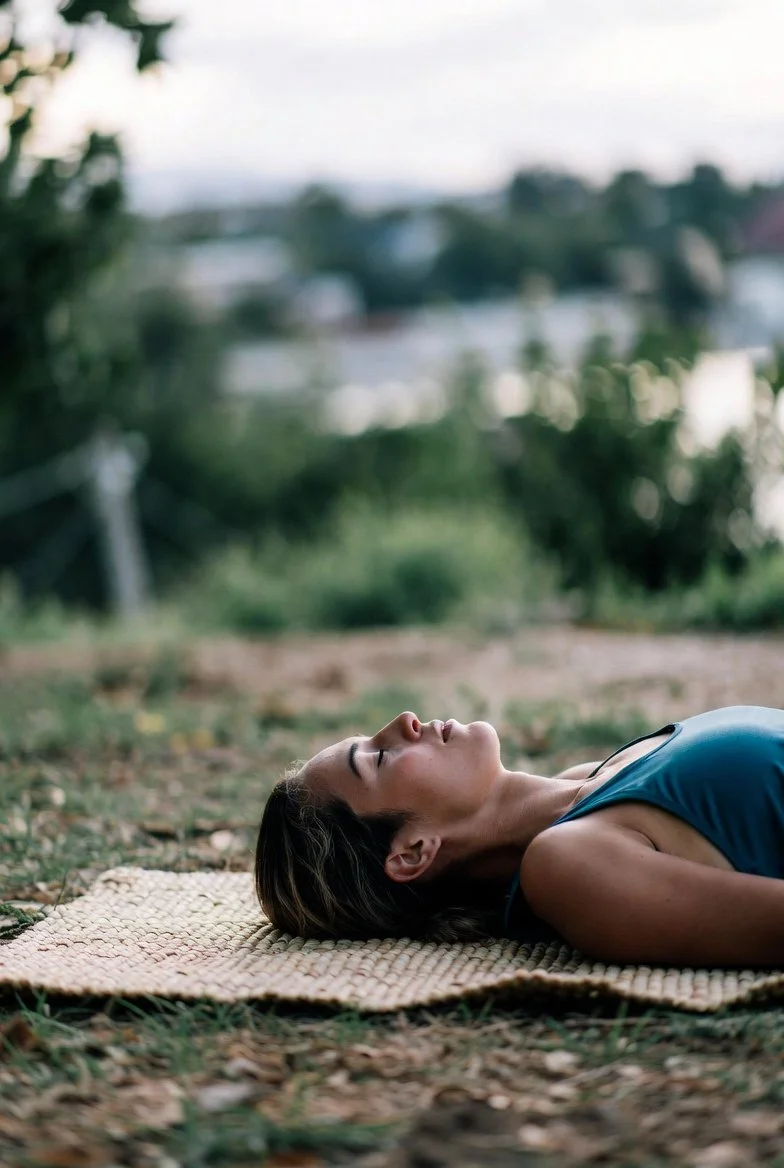 A woman with dark hair and a blue tank top lying on a woven mat outdoors, relaxing with eyes closed, in a natural setting with greenery and blurred trees or houses in the background.