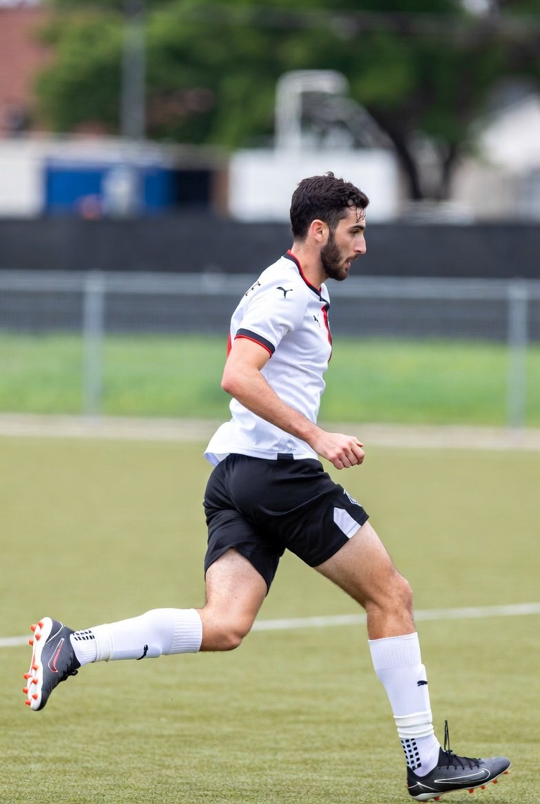 A male soccer player running on the field during a game, wearing a white shirt, black shorts, white socks, and black cleats.