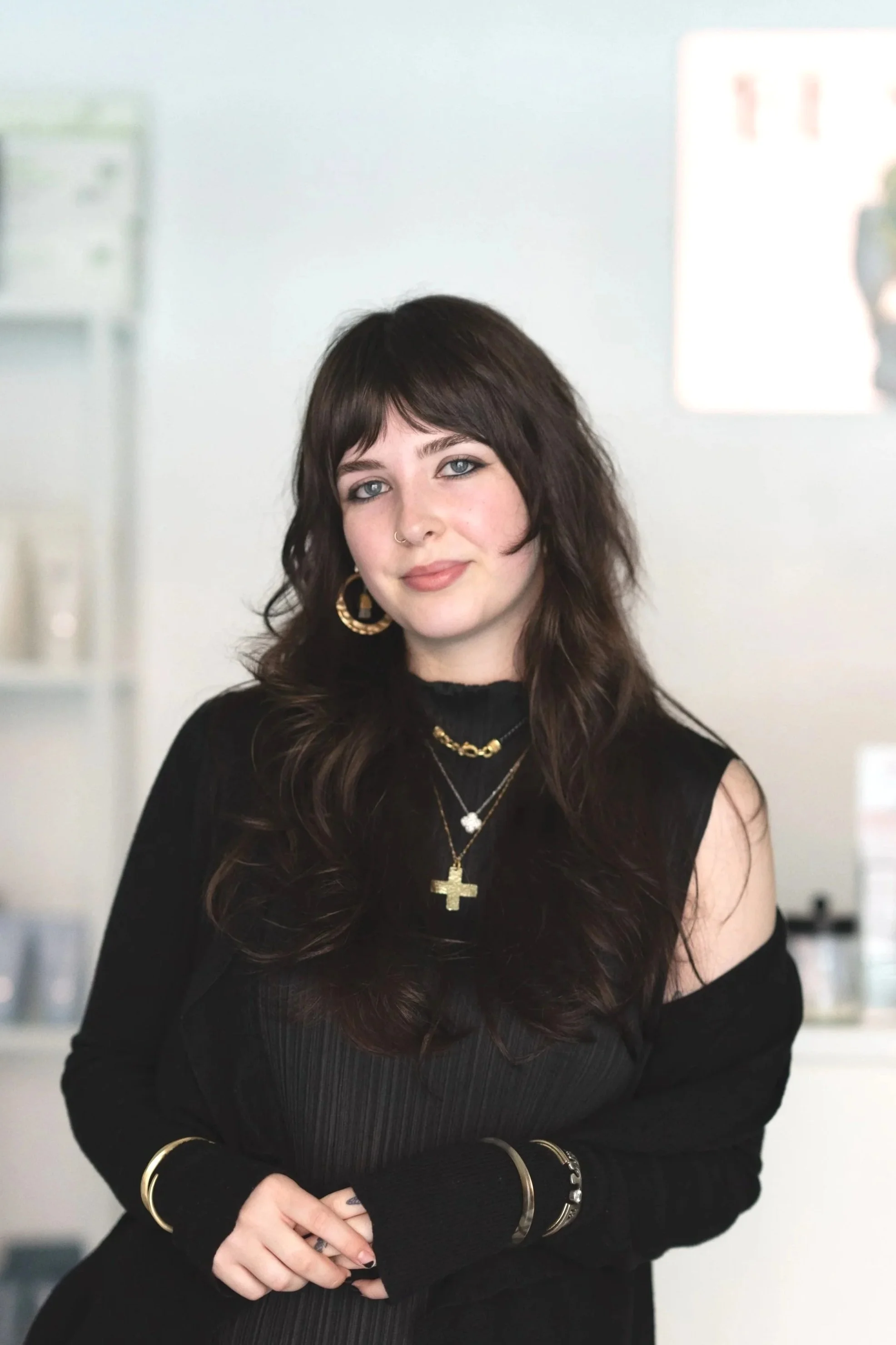 A young woman with shoulder-length brown hair, blue eyes, wearing a black ribbed top, layered gold necklaces, and a black outer garment, standing in front of a wall with a mural of building facades.