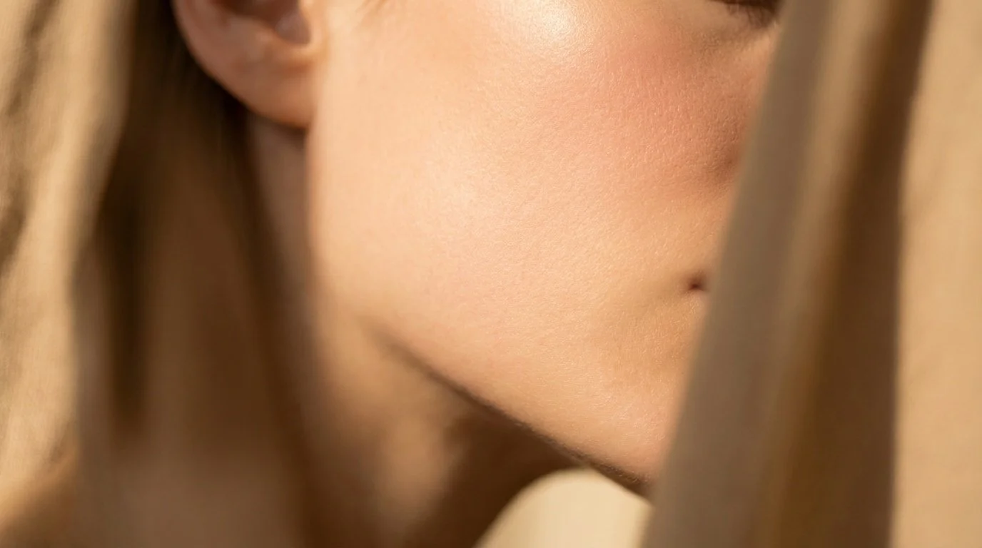 Close-up of a woman's face showing her cheek, eye, and partial nose, with smooth skin and warm lighting.