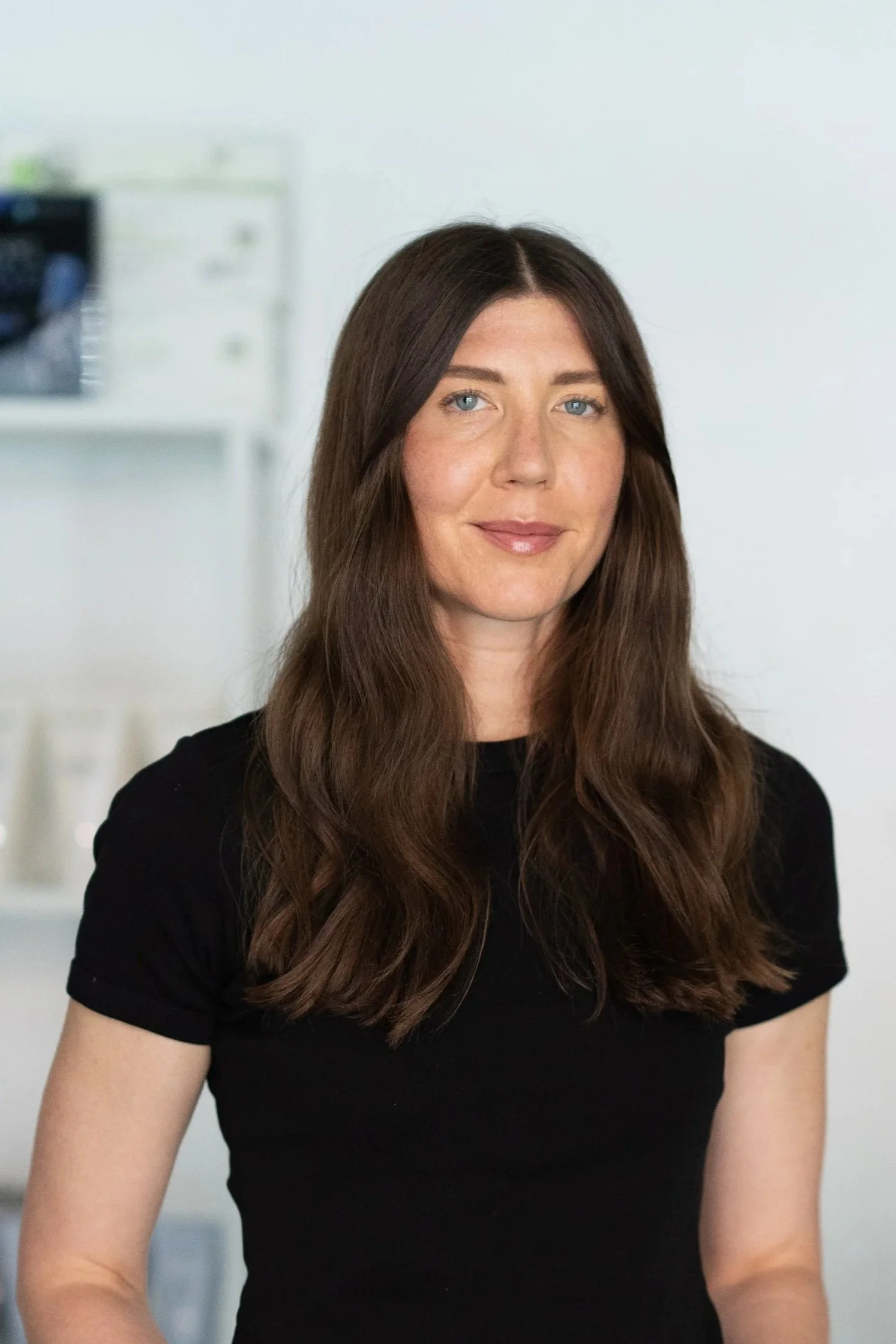 Portrait of a woman with long brown hair and blue eyes, wearing a black shirt, standing in front of a wall with a black and white mural of buildings.