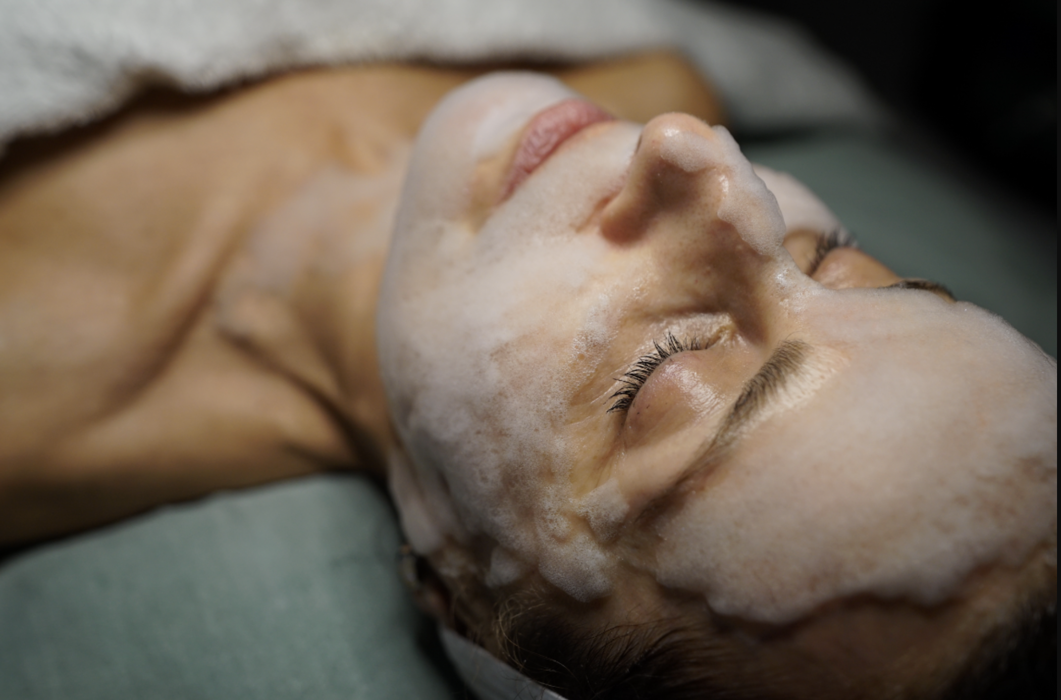 A woman receiving a facial treatment with foam on her face, lying down with eyes closed.