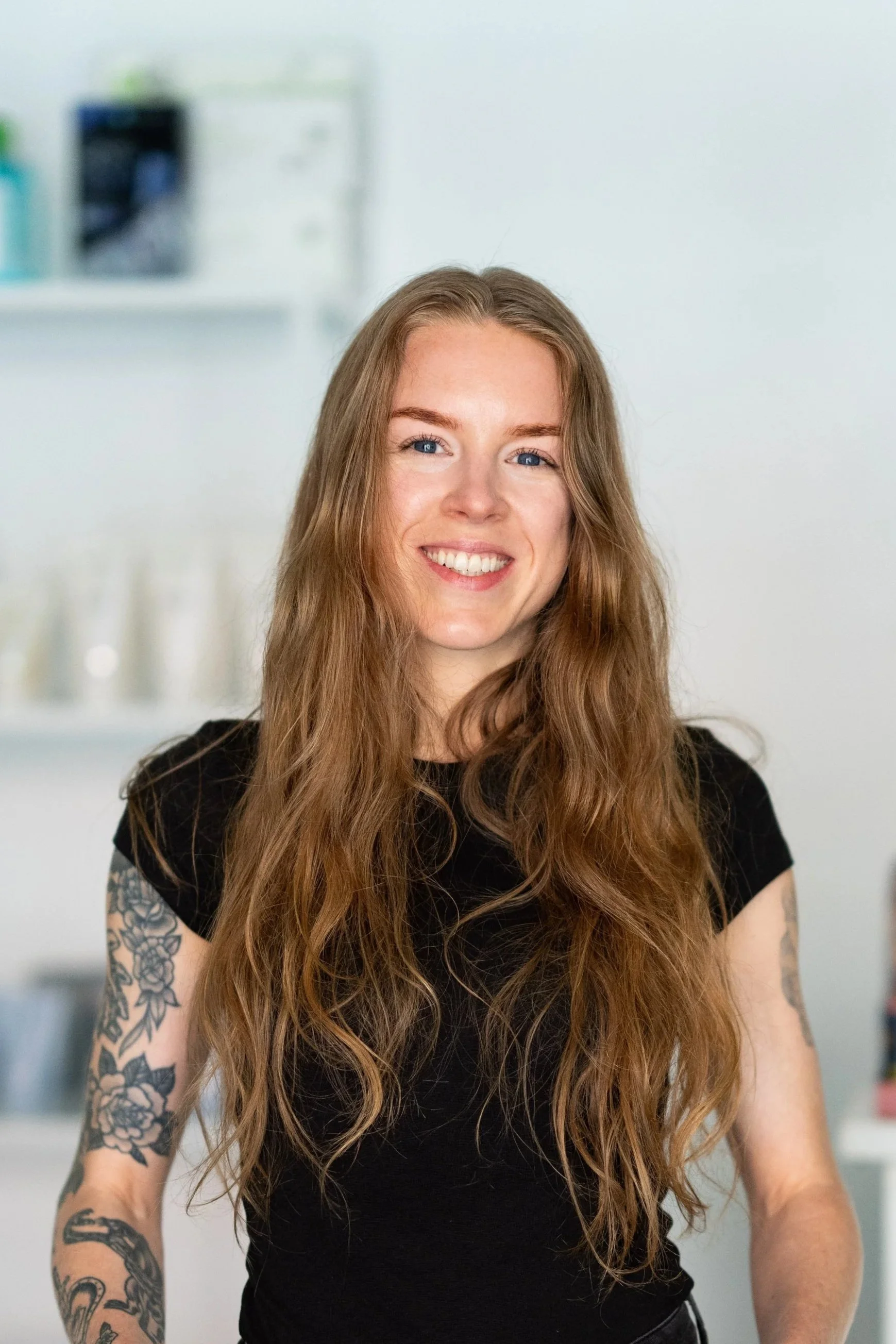 Young woman with long red hair, blue eyes, and a tattoo on her upper arm, smiling and wearing a black t-shirt, standing in front of a blurred black and white architectural background.