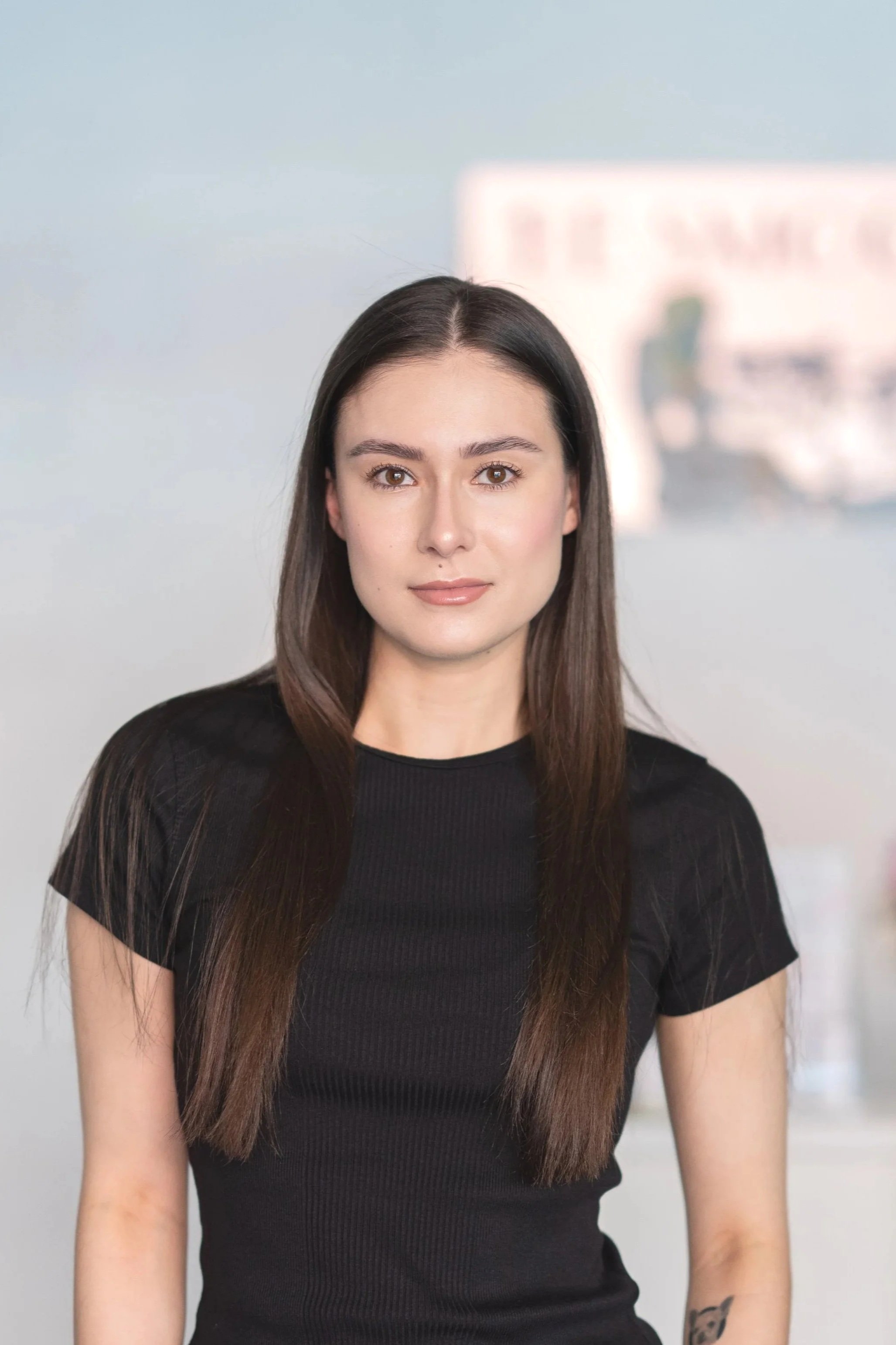 Young woman with long brown hair and light skin, wearing a black top, standing in front of a mural of a cityscape.