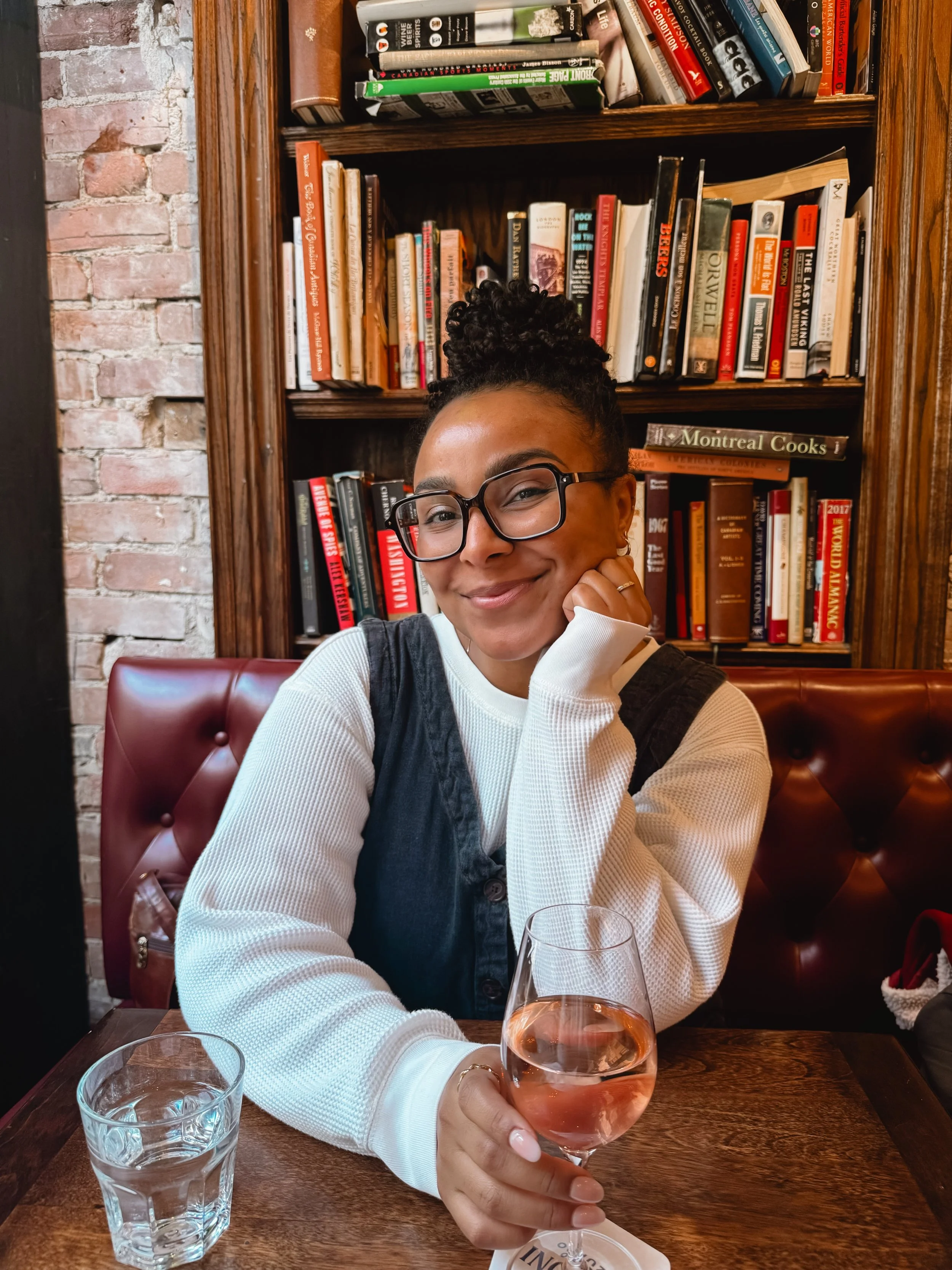 A young woman with glasses smiling and holding a glass of rosé wine. She is sitting at a wooden table in a cozy restaurant with exposed brick and wooden bookshelves behind her filled with books. There is also a glass of water on the table.