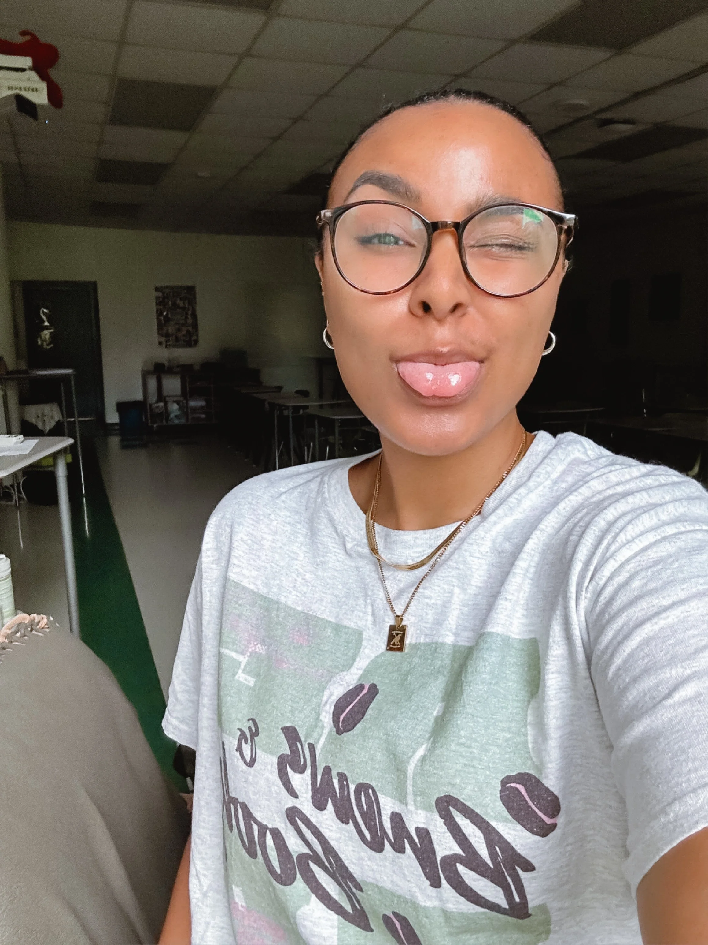 A person taking a selfie in a room, wearing glasses, a white T-shirt with colorful text and graphics, and displaying a playful expression with tongue sticking out. The room has a ceiling with some ceiling tiles missing, a dark door, and tables and chairs in the background.