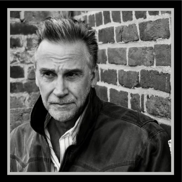 A black and white photo of an older man with gray hair and a beard, wearing a dark jacket over a light-colored collared shirt, standing outdoors against a brick wall.
