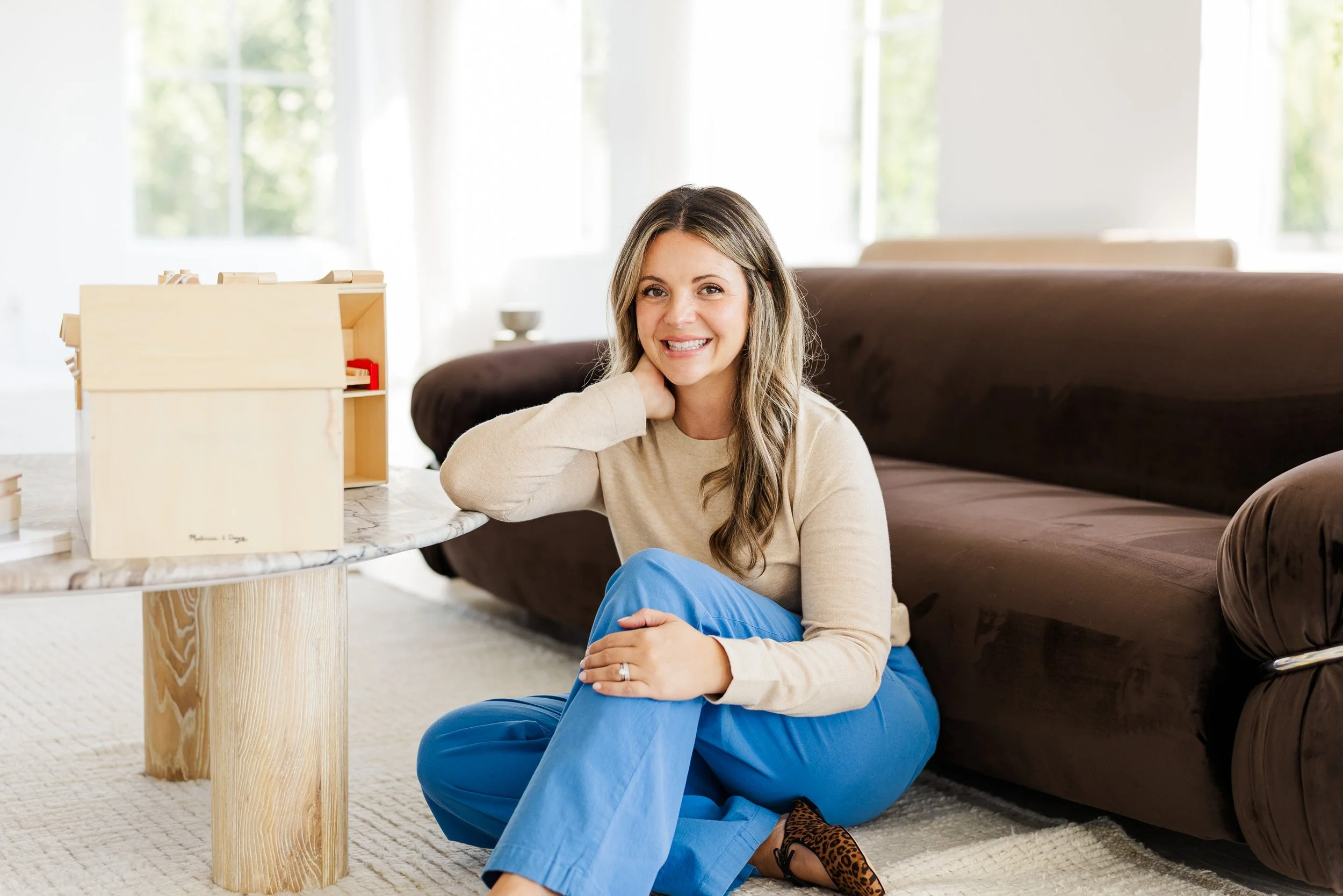A woman with long brown hair, wearing a beige long-sleeve shirt and blue pants, sitting on the floor next to a coffee table in a bright living room with large windows. She is smiling, resting her chin on her hand, and has a leopard print shoe visible. There is a dark brown sofa and a wooden box or shelf on the table.