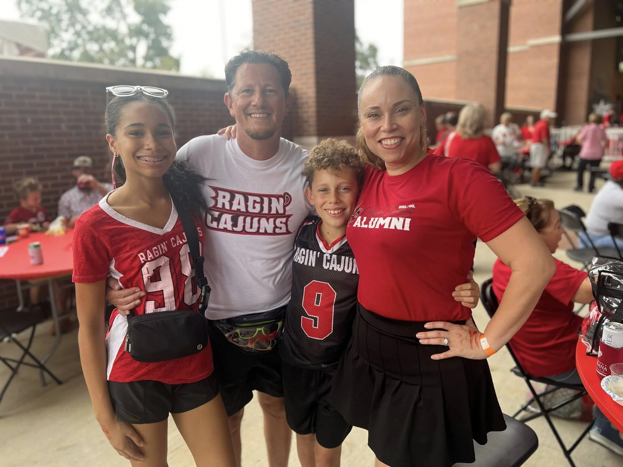 Group of four smiling people at a sports event, wearing Ragin Cajuns apparel, with a brick wall and other attendees in the background.