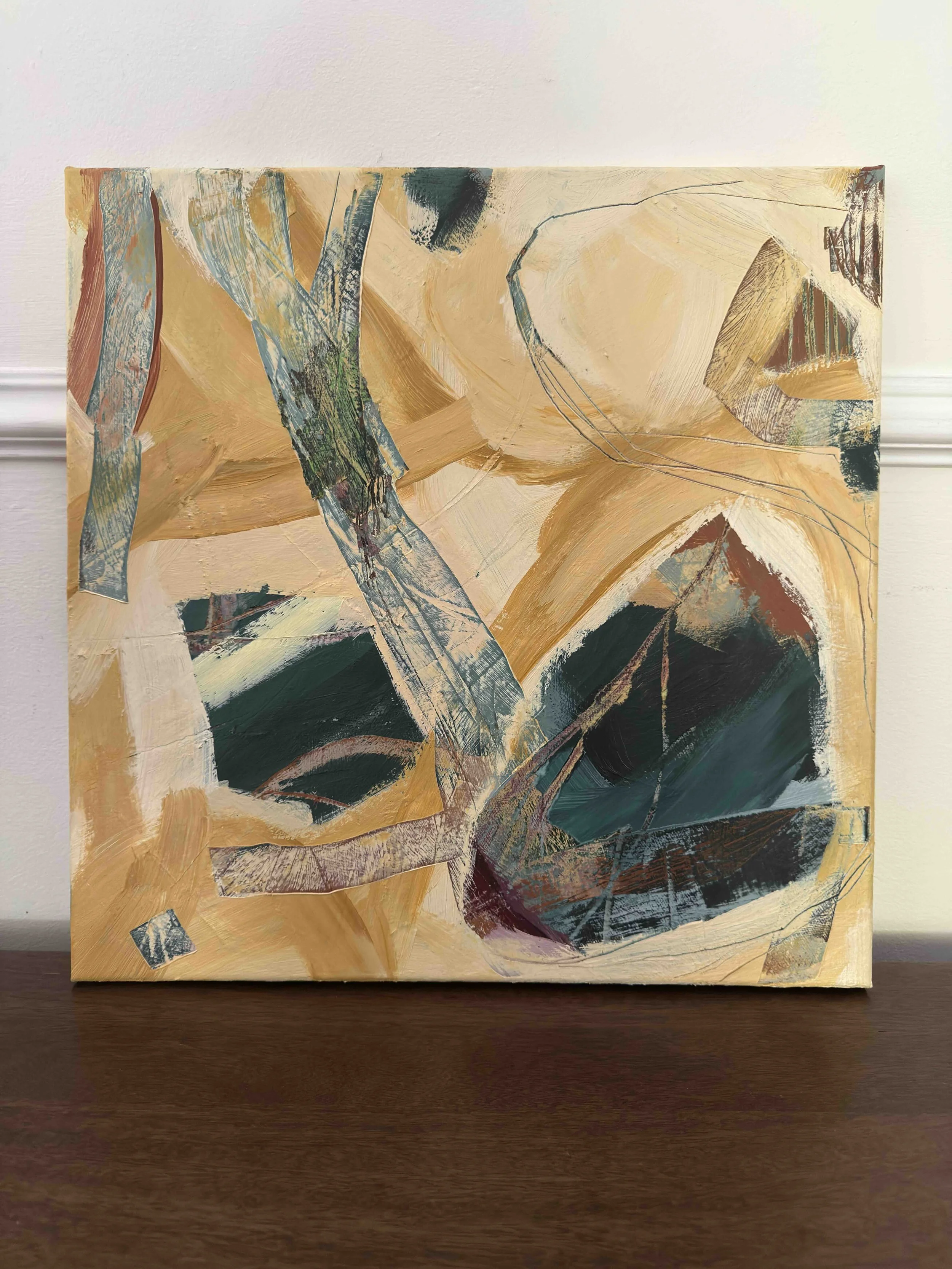 Abstract painting with tan, dark green, blue, and black brush strokes and shapes, mounted on a wall above a wooden surface.