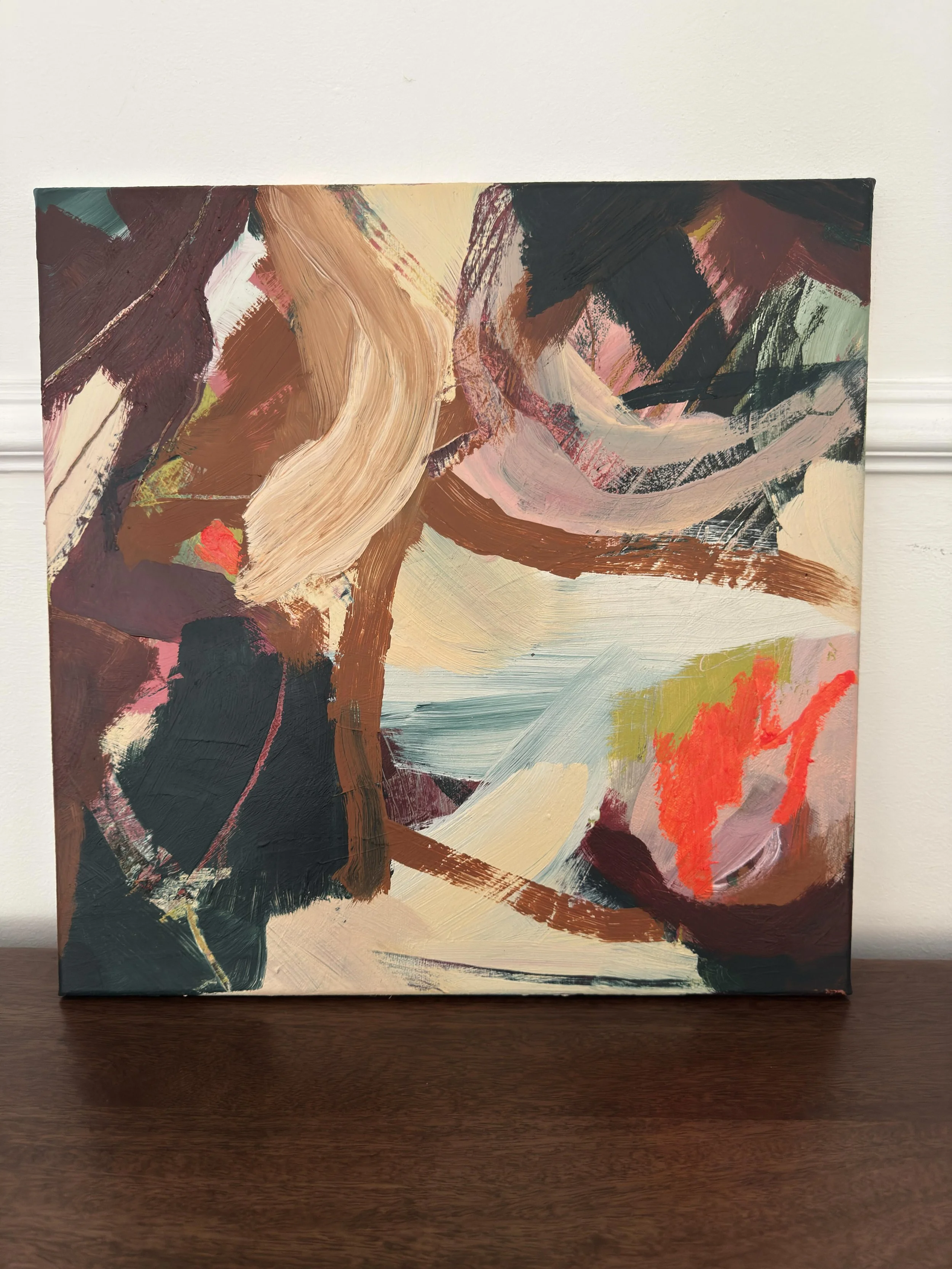 Abstract painting with sweeping brushstrokes of beige, pink, black, brown, and red on a canvas, resting on a dark wooden surface.