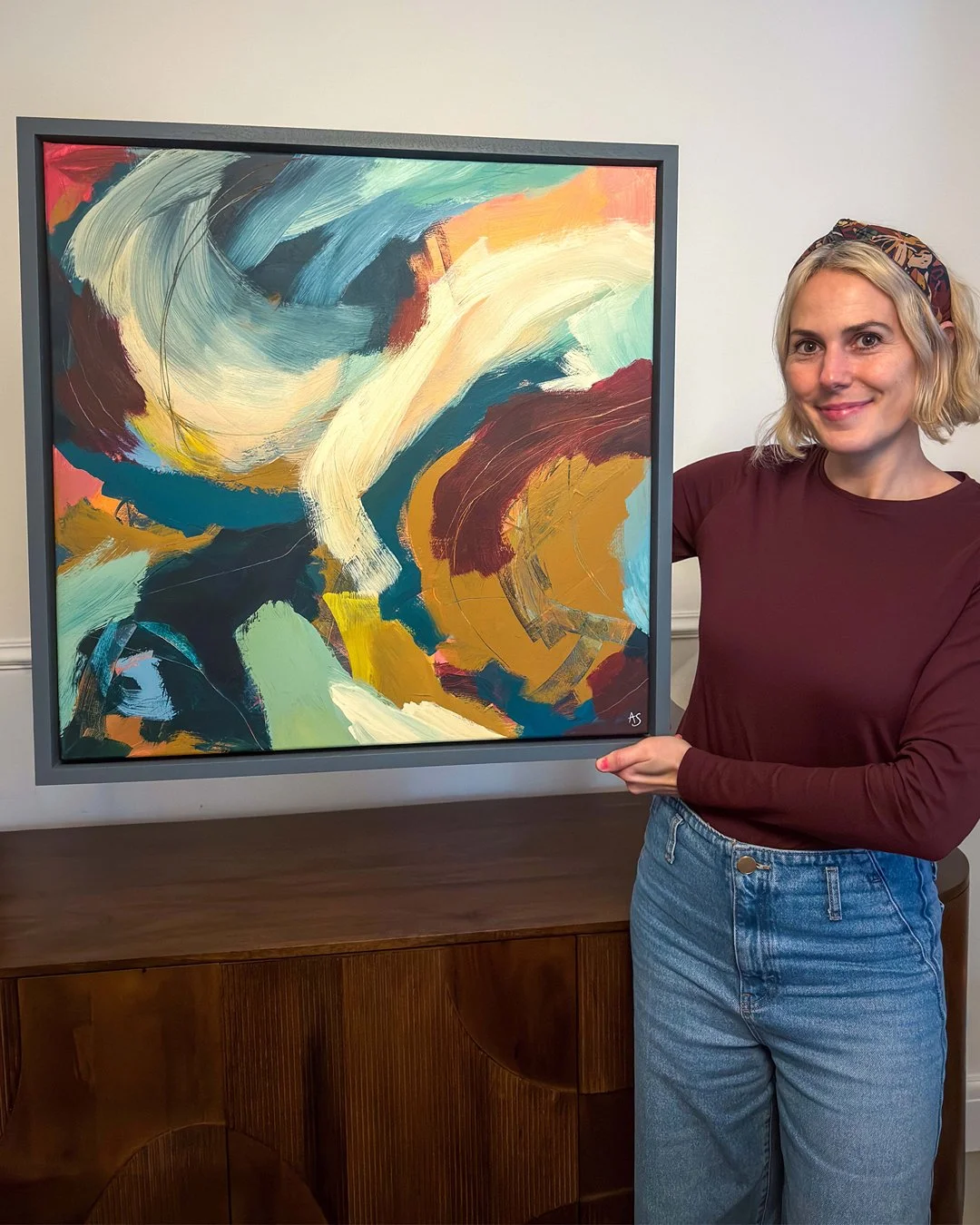 Woman with blonde hair, wearing a maroon shirt and blue jeans, standing next to an abstract painting with bold brushstrokes and vibrant colors.