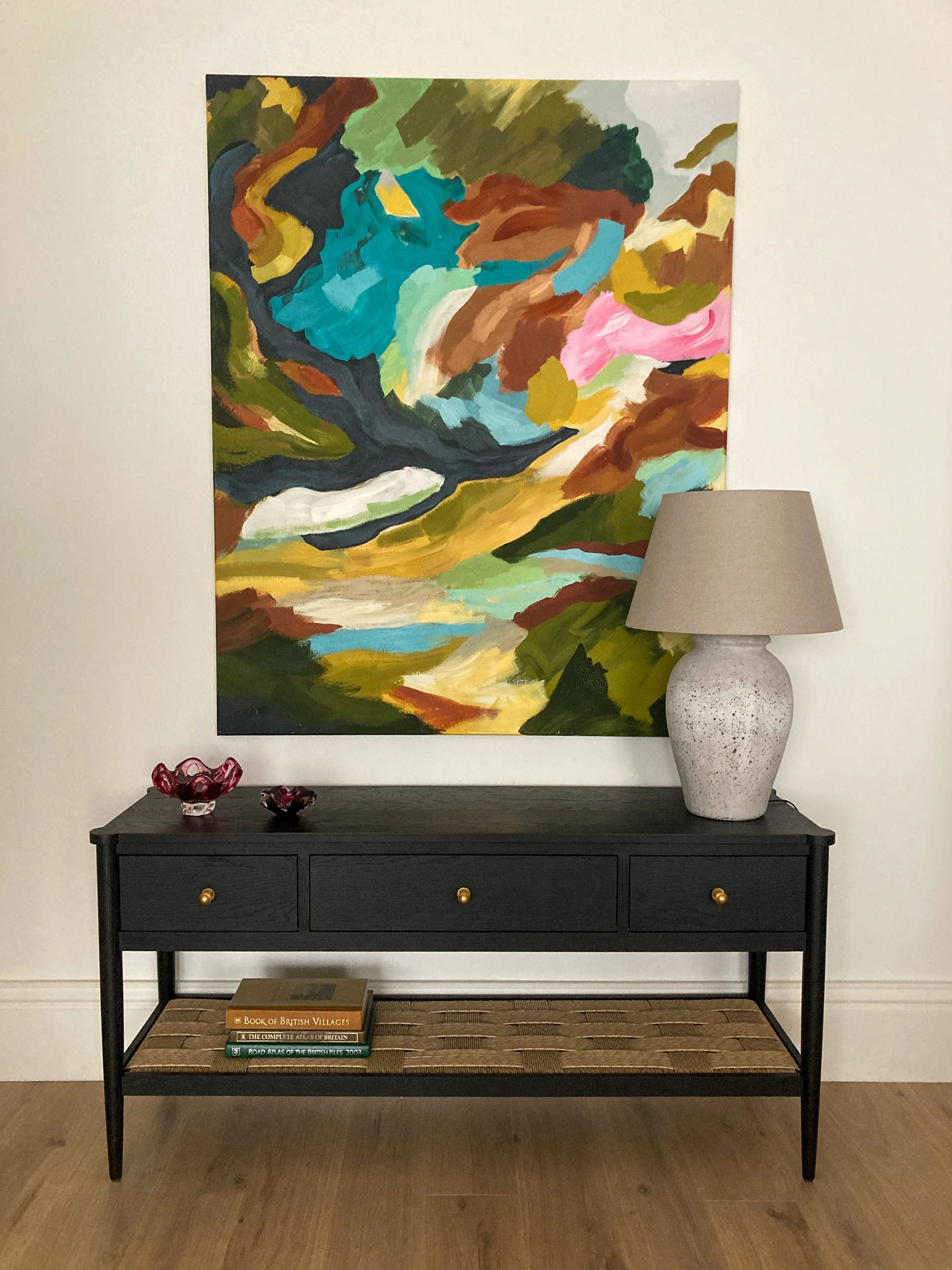 A black console table with three drawers with gold knobs, a woven shelf, and books on the lower shelf. Two decorative bowls on the left, a large white textured ceramic lamp with a beige shade on the right. A large colorful abstract painting hangs behind the table. The setting is against a white wall with wood flooring.