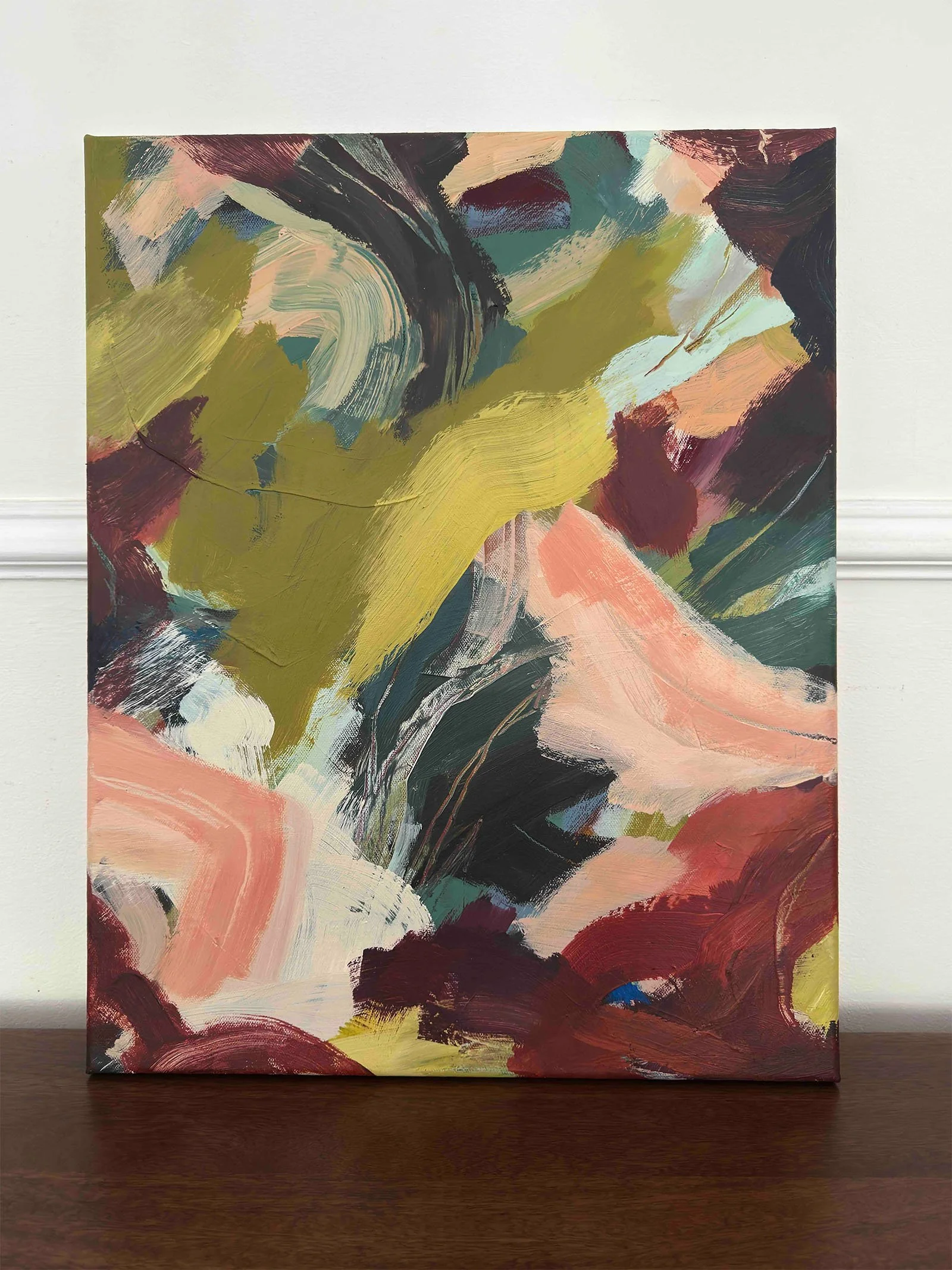 Abstract painting with bold brushstrokes in hues of green, pink, black, and white, displayed on a wooden surface against a white wall.