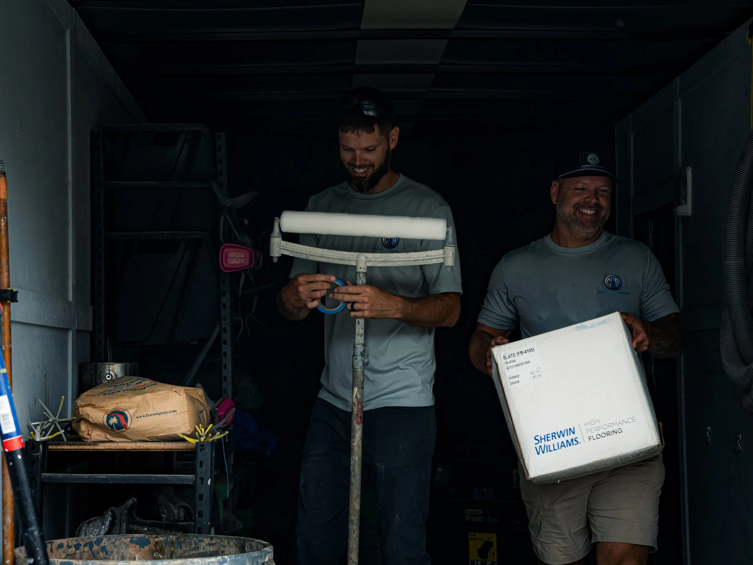 Two men working in a garage or storage area, one holding a box labeled Sherwin Williams high performance flooring, both smiling.