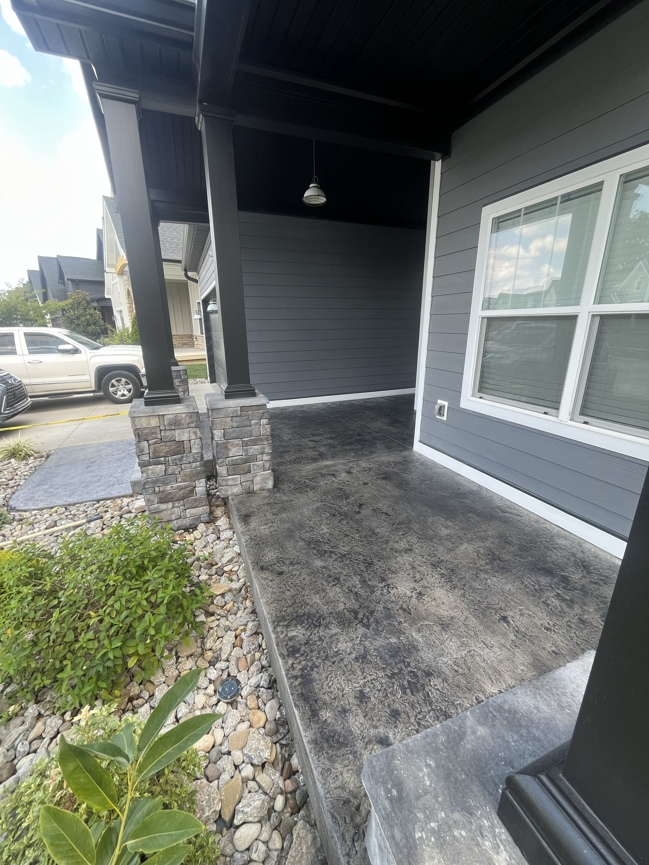 Clean front porch with dark colored concrete floor, attached to a house with grey siding, large window, and decorative brick pillars supporting the porch roof. Small plants and rocks in garden bed nearby, parked cars visible in background.