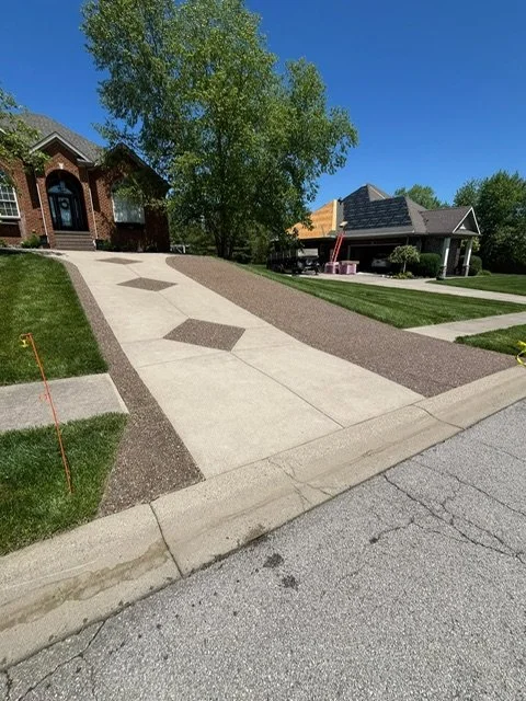 Concrete driveway with decorative inlay pattern leading to a brick house and a neighboring house with a porch, trees, grass, and a clear blue sky.