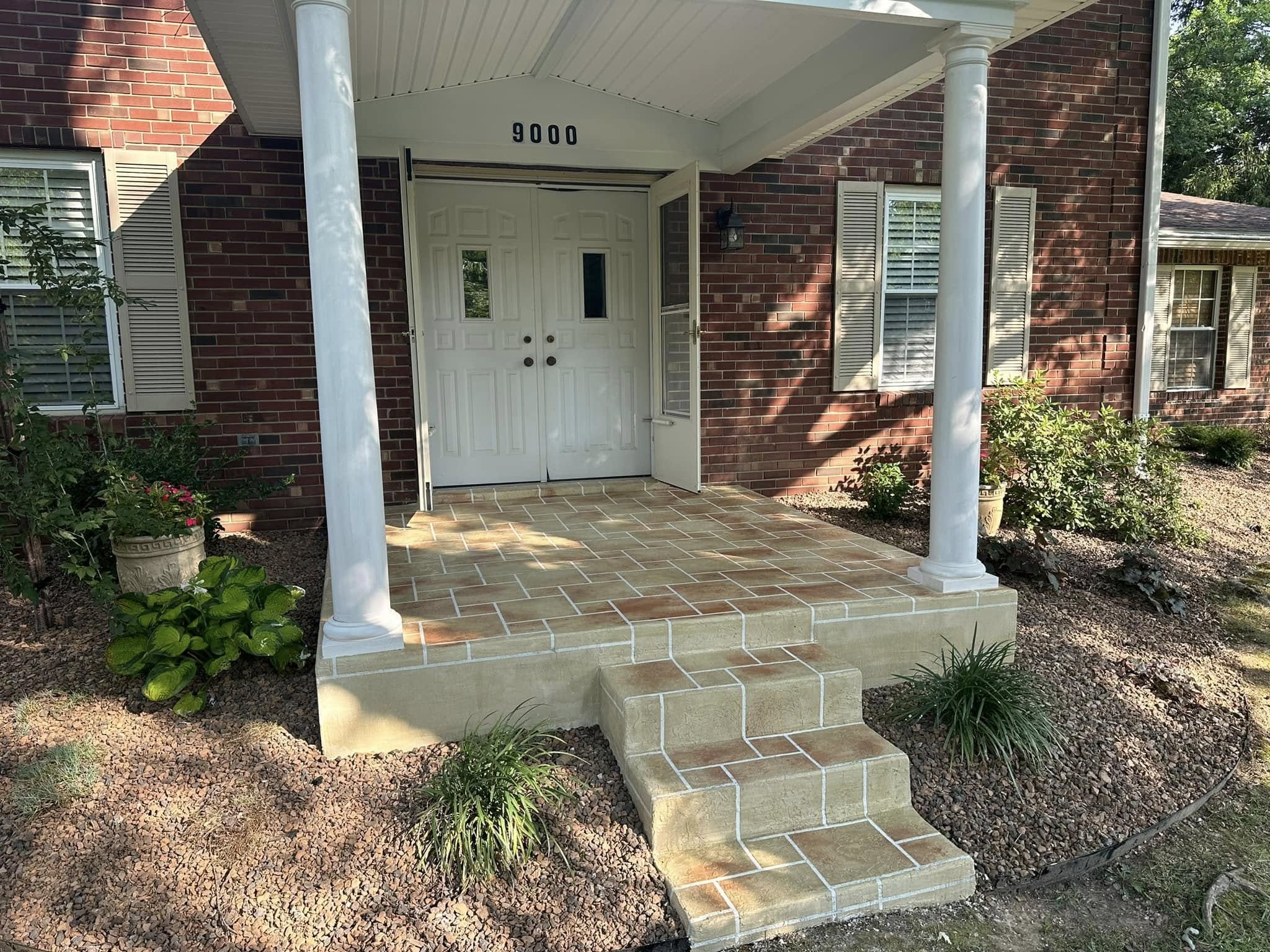 Front porch of a brick house with a white double door entrance, four steps leading up, supported by two white round columns, with greenery and plants on either side.