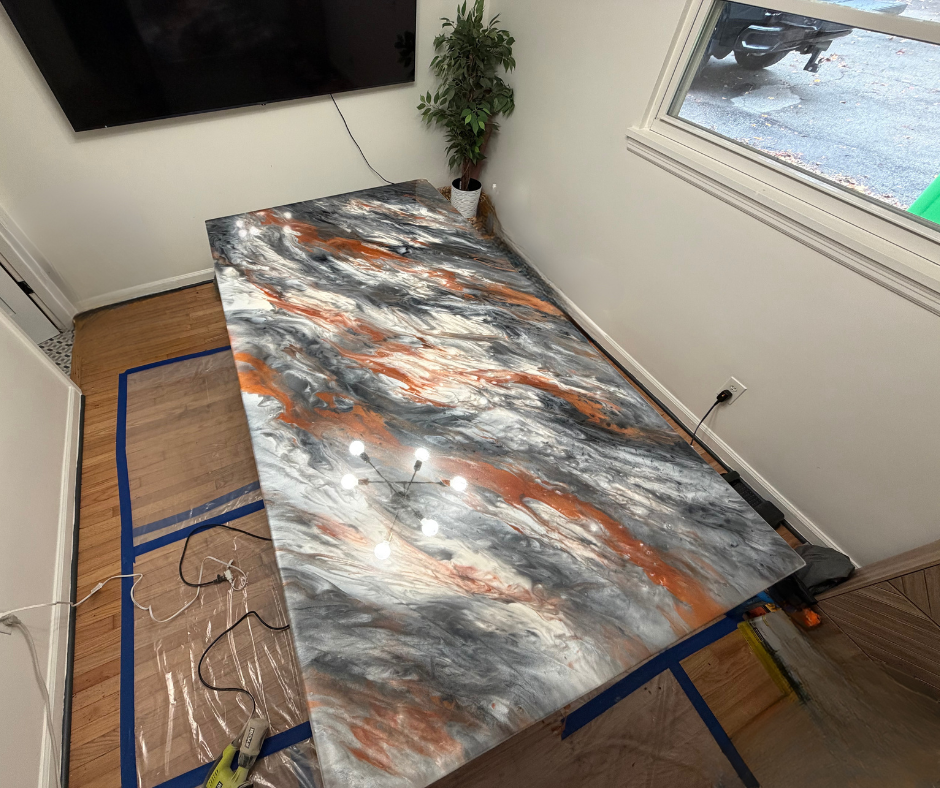 A large abstract painting with swirls of black, white, gray, and orange on a wooden table in a room with hardwood floors, a flat-screen TV on the wall, a potted plant, and a window showing an outdoor driveway and a green trash bin.
