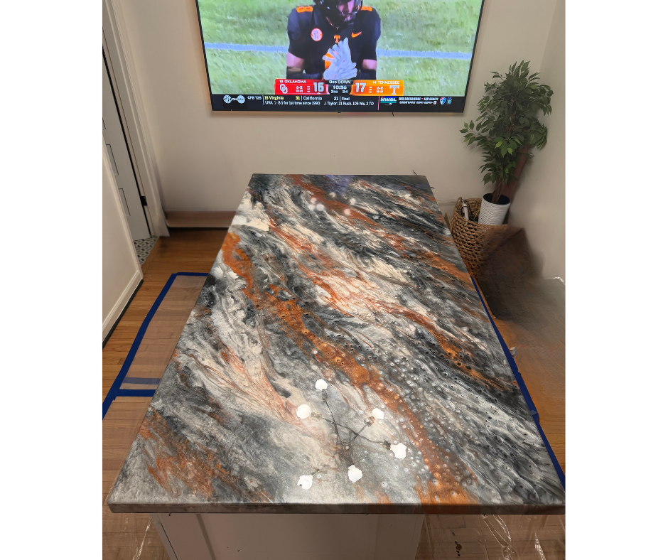 A marble countertop with black, white, gray, and brown swirls in a kitchen, with a potted plant in a basket in the corner and a TV mounted on the wall showing a football game.
