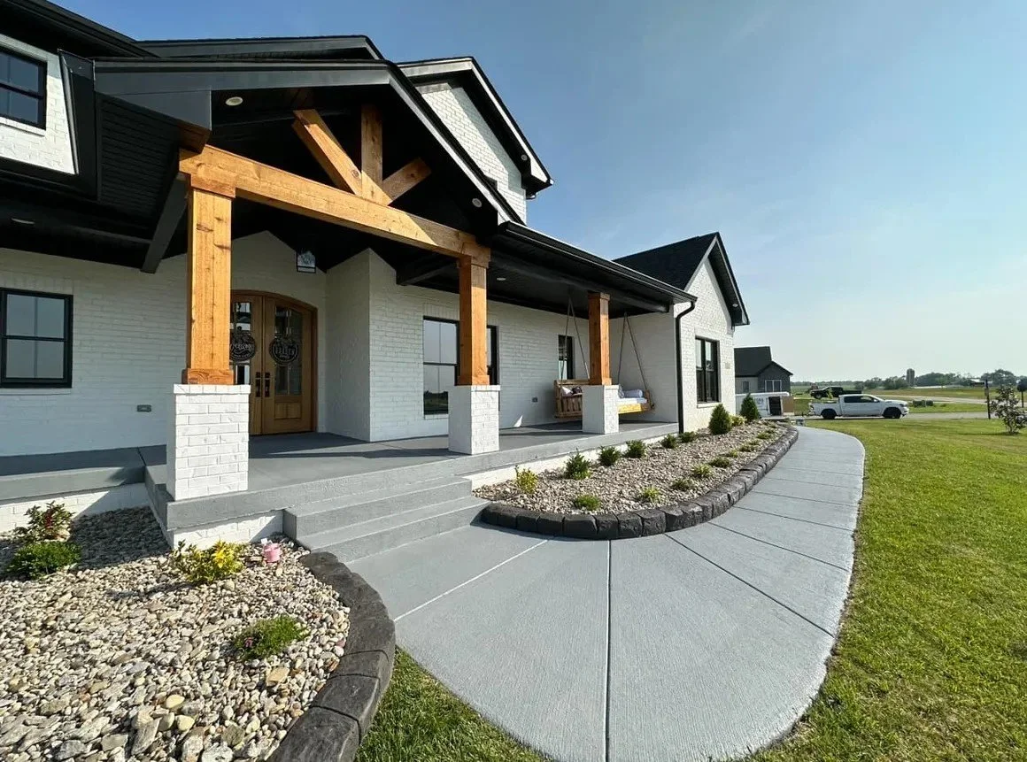 Exterior view of a modern house with a white brick facade, a covered porch with wooden beams, a porch swing, landscaped garden beds, and a concrete walkway, with a car parked in the background under a clear sky.