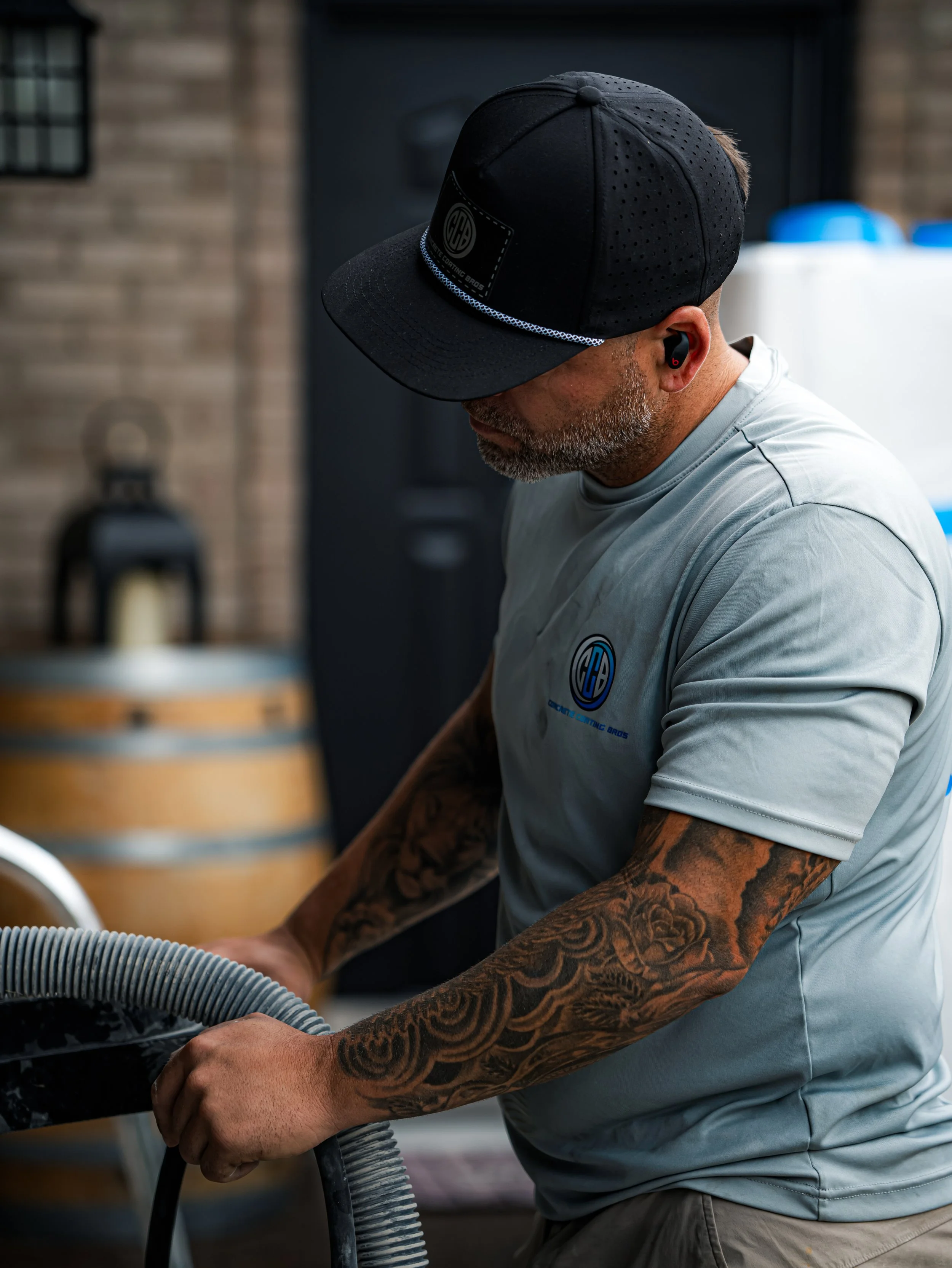 A man with tattoos on his arms, wearing a gray T-shirt, a black cap, and wireless earbuds, is working with a hose.