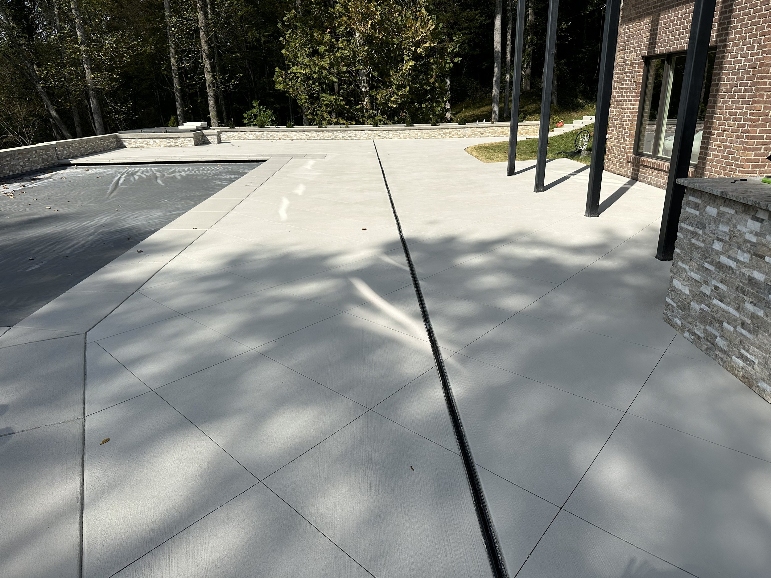 Newly constructed spacious backyard patio with white concrete tiles, adjacent to a brick house with large glass doors, surrounded by trees and greenery in the background.