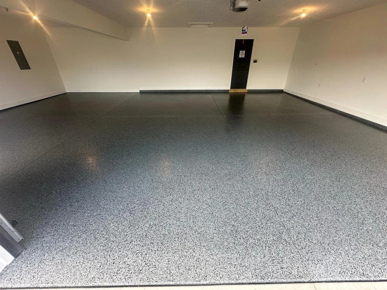 Empty room with speckled grey epoxy flooring, white walls, and a ceiling with recessed lighting. There is a black door at the back with a small sign or paper attached and an overhead garage door opener unit.