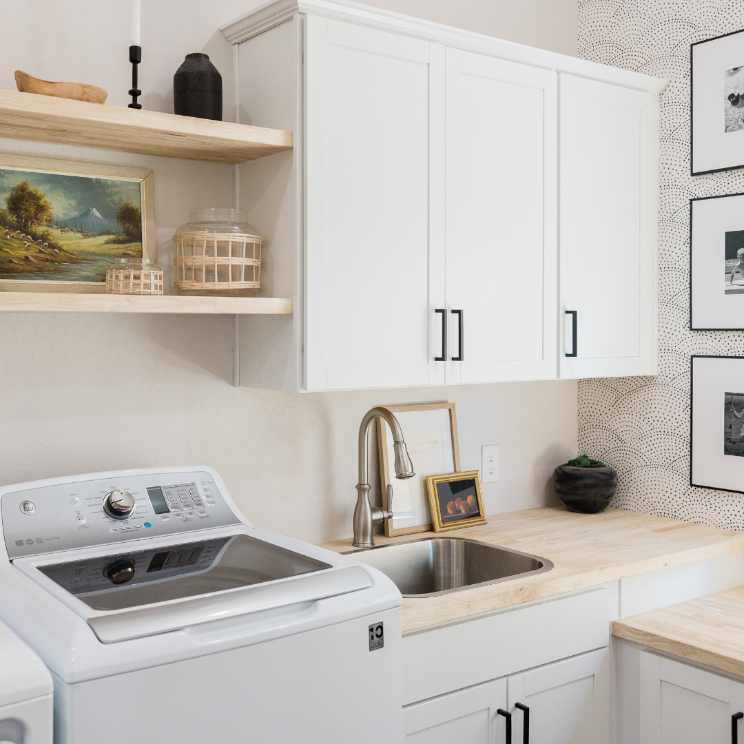 Laundry room with white cabinets, wooden countertops, a washing machine, a kitchen sink with a bronze faucet, and decorative items including framed photos, a black pot, and wall art.