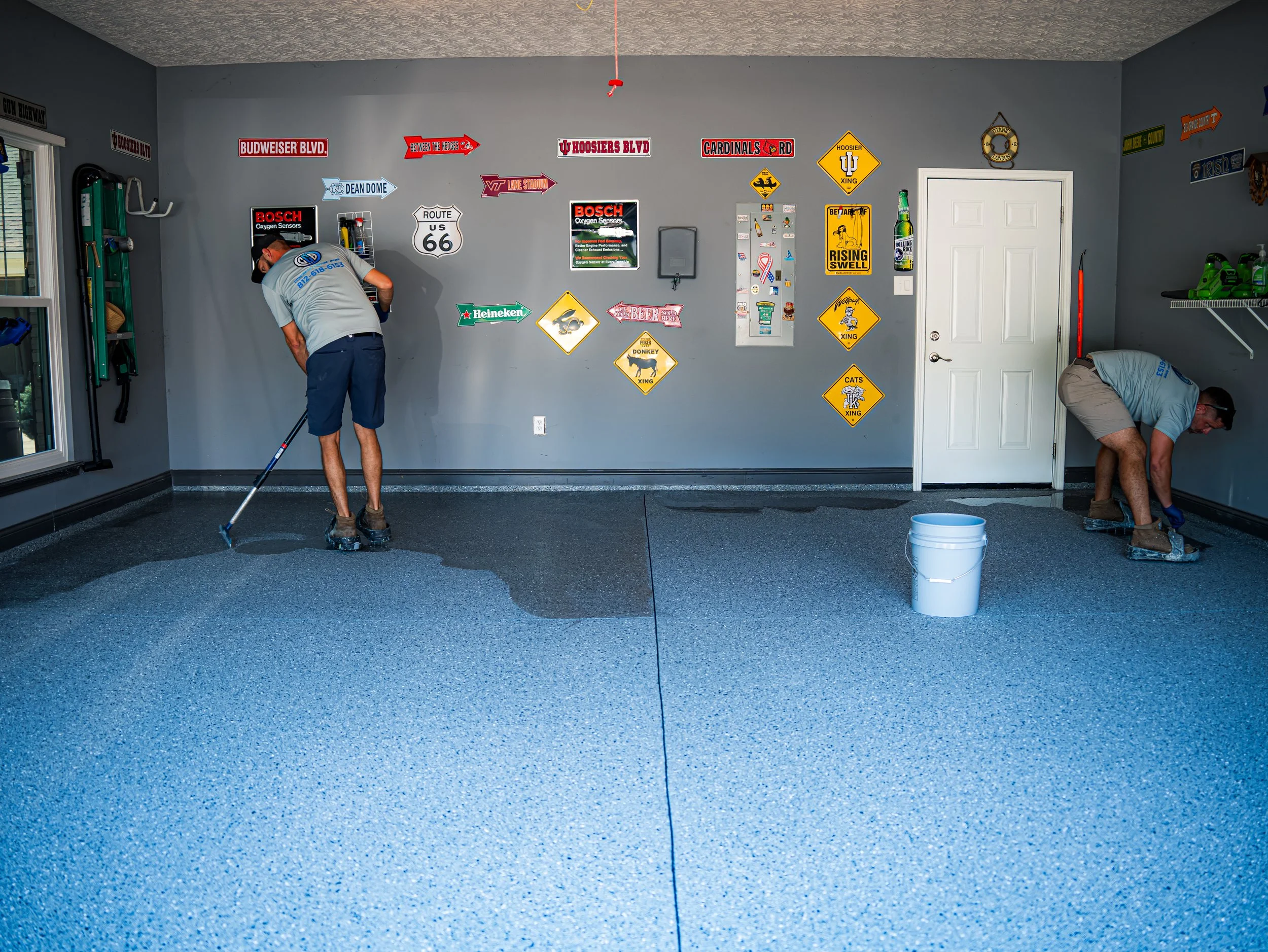 Two men are installing epoxy flooring in a garage, with one applying a coat of epoxy on the left side and the other on the right side. The garage walls are decorated with sports and traffic signs, and there are shelves with green bottles on the right.