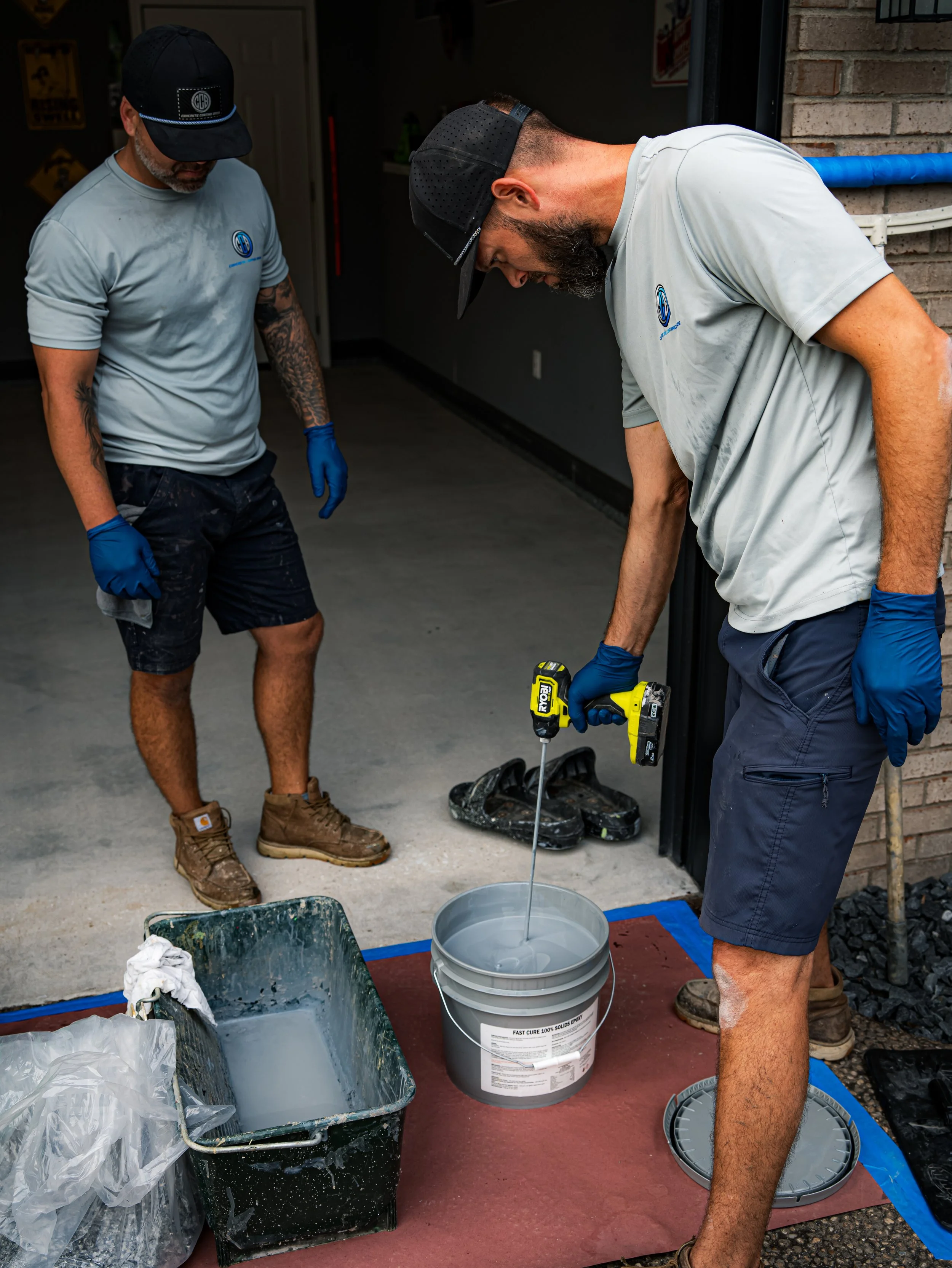 Two men working in a garage, one pouring liquid from a yellow power drill into a bucket, the other observing, with tools and safety gear nearby.