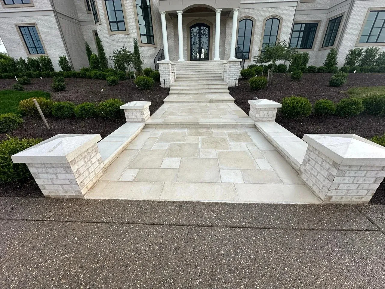 Front entrance of a large white brick house with stone stairs, columns, and landscaped garden with bushes.