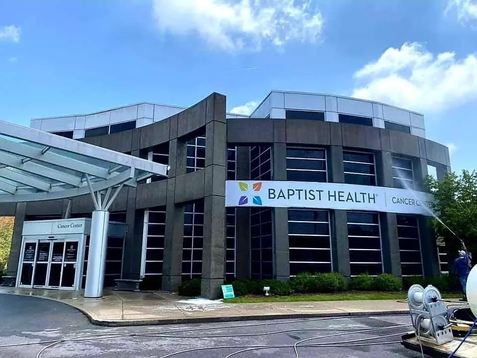 Exterior of Baptist Health Cancer Center building with a large sign, glass entrance, and fire hoses on the ground, under a partly cloudy sky.