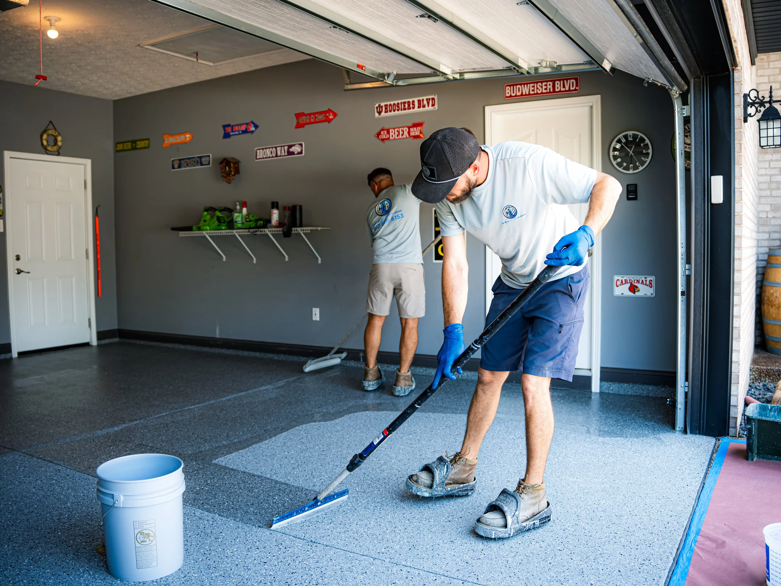 Two men working on installing or cleaning a garage floor. The man in the foreground is using a floor buffer pad, wearing gloves and sandals with socks, and the man in the background is using a smaller tool, both wearing gray shirts with a logo.