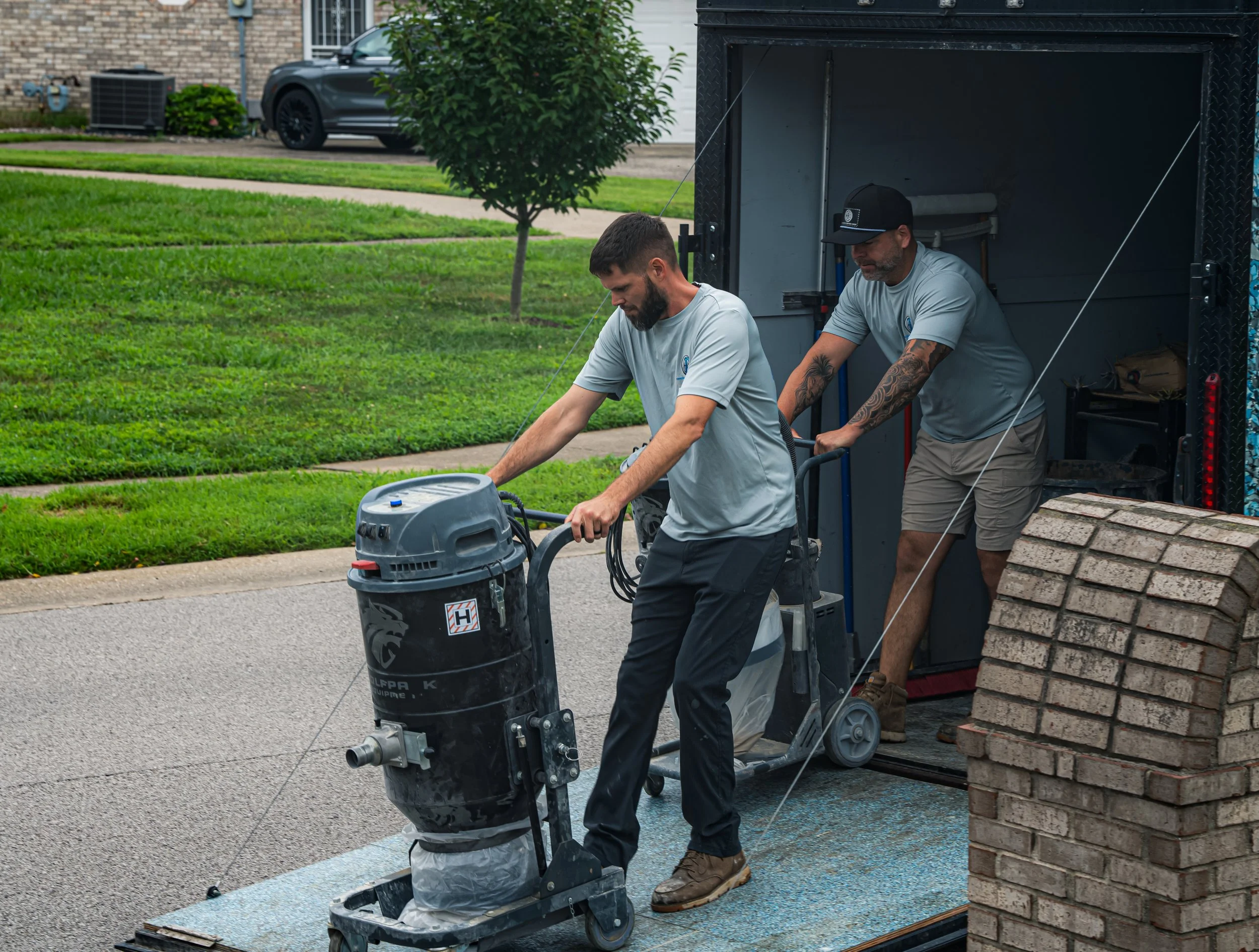 Two men working on a construction project on a residential street, with a truck, grass, a tree, and a parked car in the background.