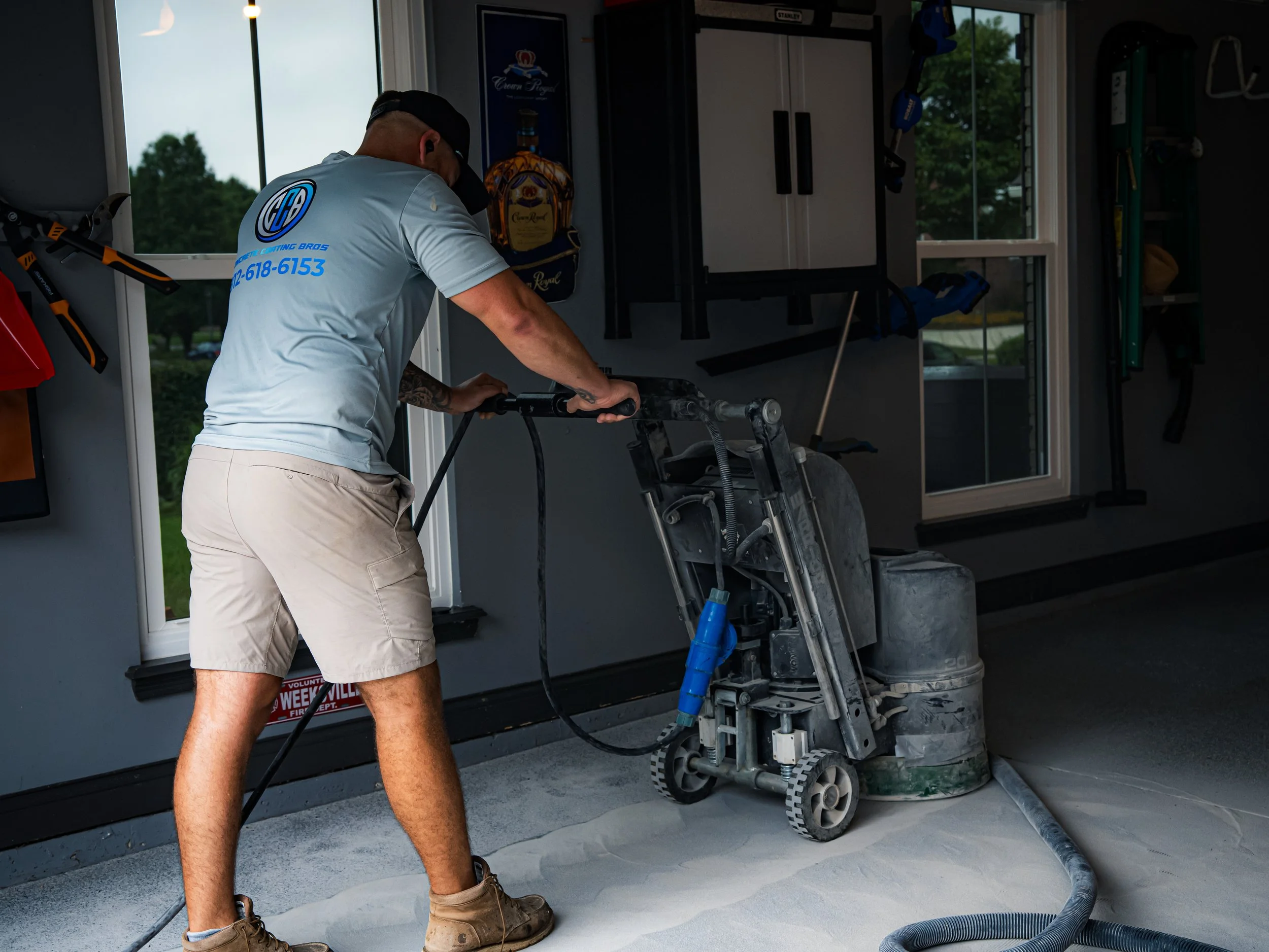 A worker using a concrete grinding machine on a floor inside a garage with tools and cabinets visible in the background.