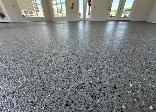 Empty room with newly installed speckled gray basement or garage floor, large windows, and a view of buildings outside.