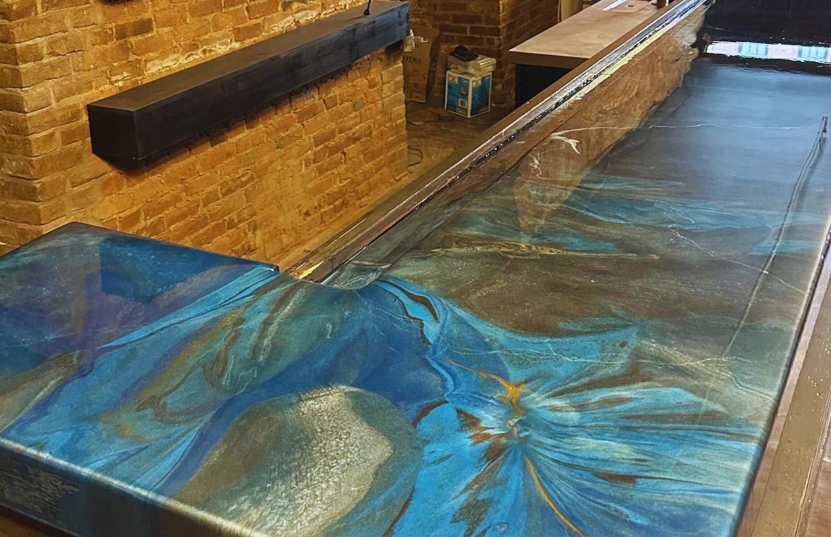 A long table with a colorful abstract design on its surface, featuring blue, brown, green, and gold swirls. The background shows a brick wall and some boxes and furniture.