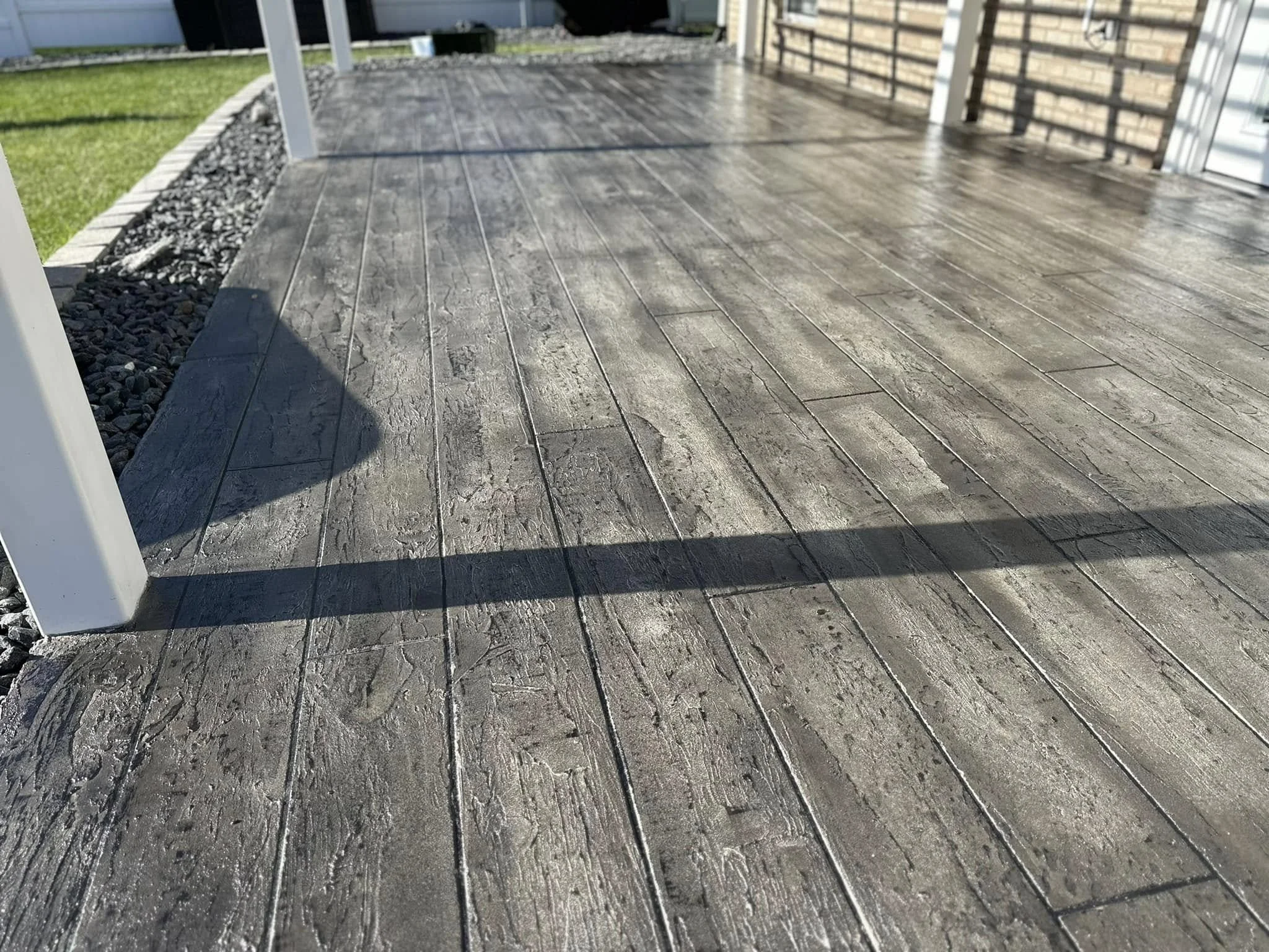 A wooden deck on a porch with a shadow cast over part of it, adjacent to a grassy yard and a small border of gravel or rocks.